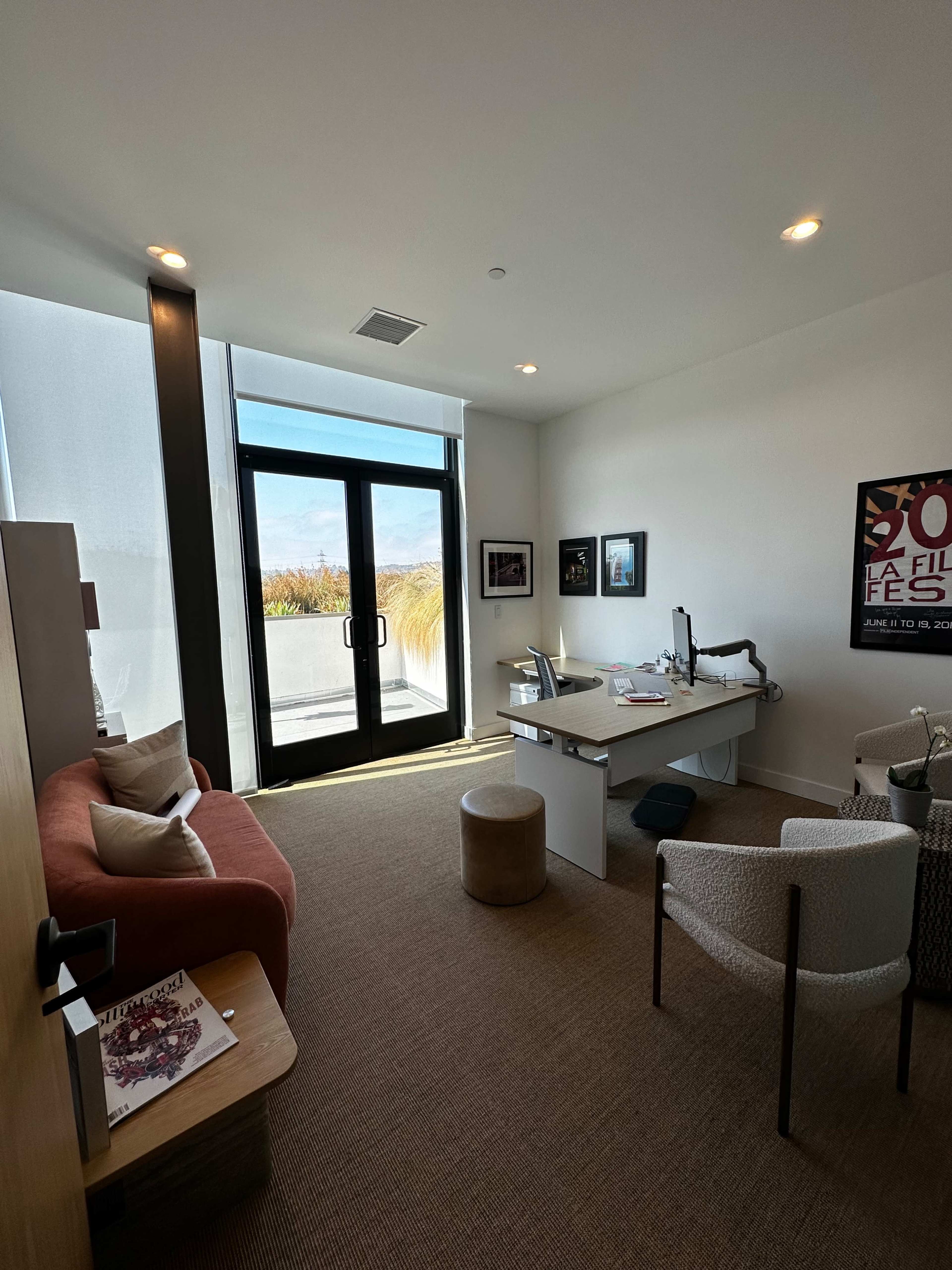 The image shows a modern office space with a desk, two chairs, a sofa, and large windows overlooking a balcony.