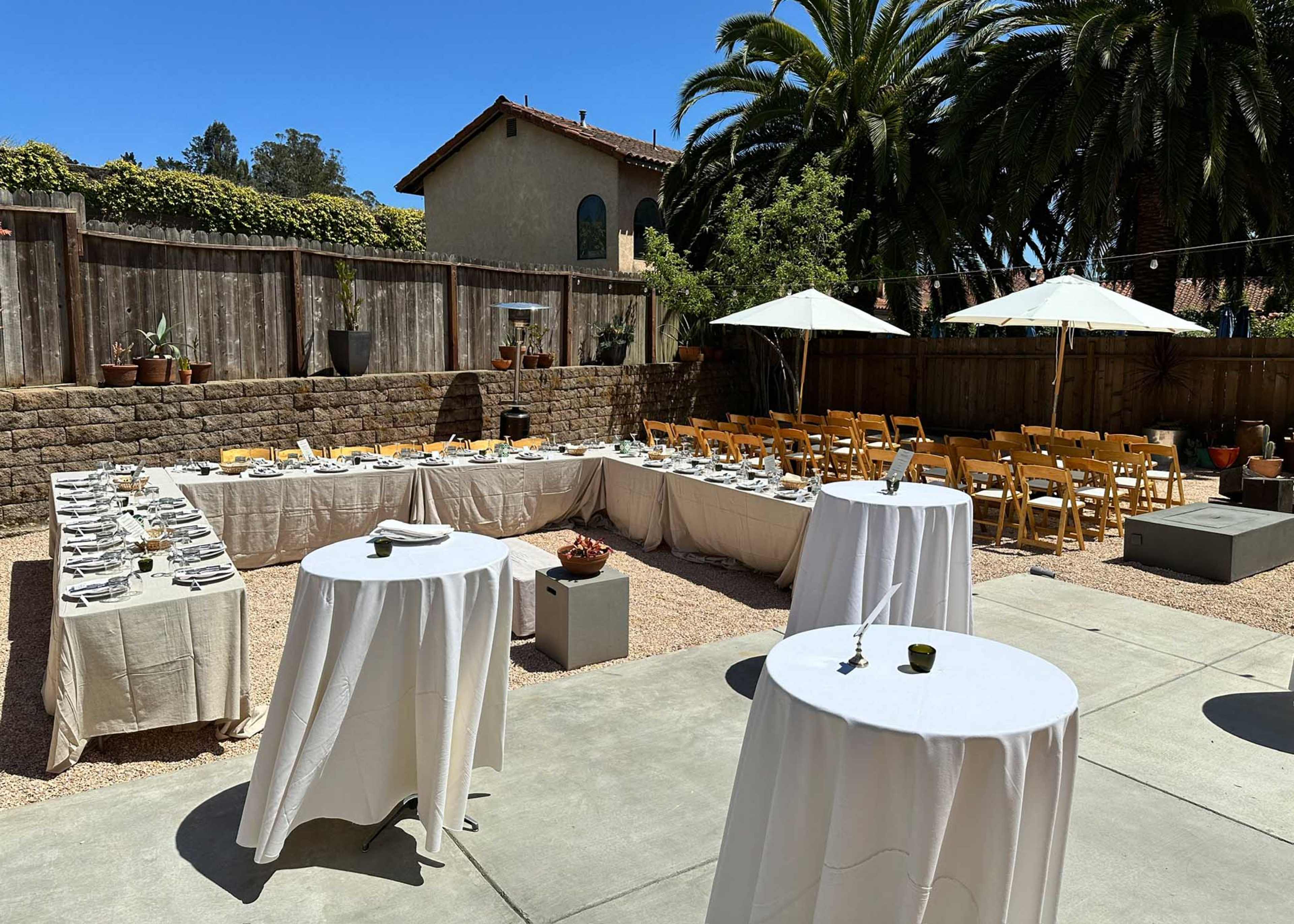 A well-arranged outdoor event space features several tables set for dining, surrounded by palm trees and umbrellas, with additional seating in the background.