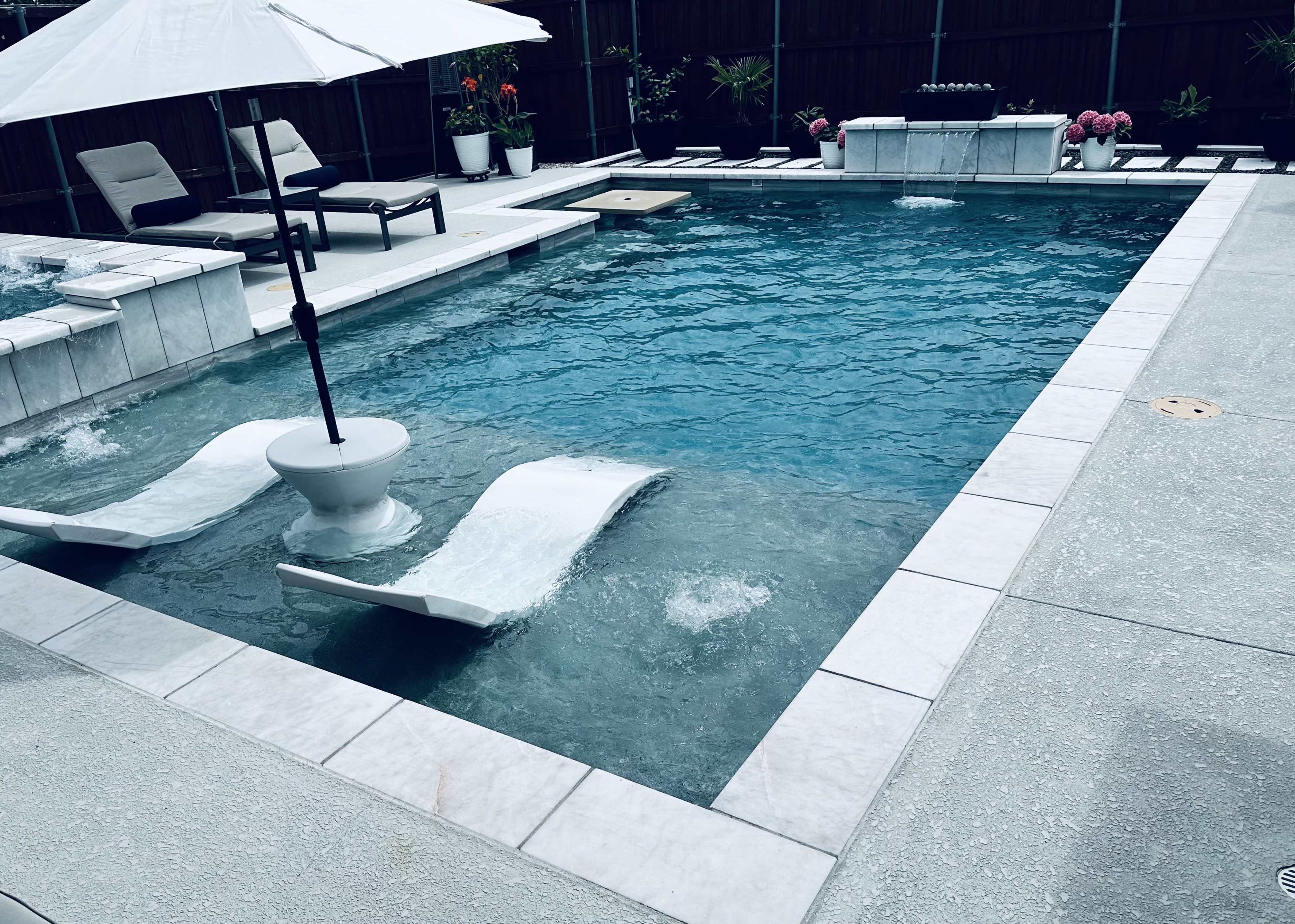 The image shows a rectangular swimming pool with two lounge chairs partially submerged in water, surrounded by a patio area with decorative plants and an umbrella.