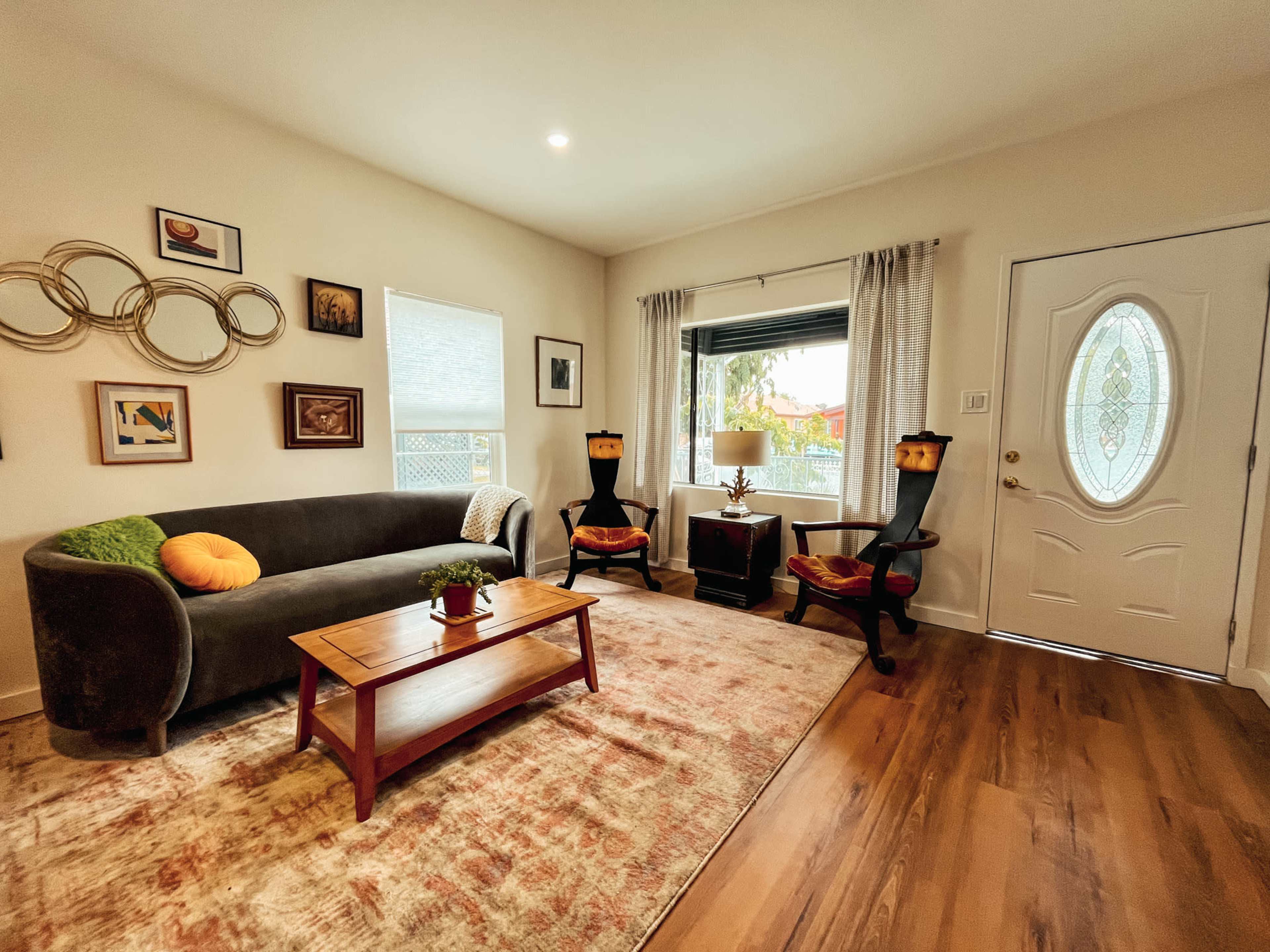 The image shows a living room with a gray sofa, two dark chairs, a wooden coffee table, and a door leading outside, complemented by framed art on the walls.