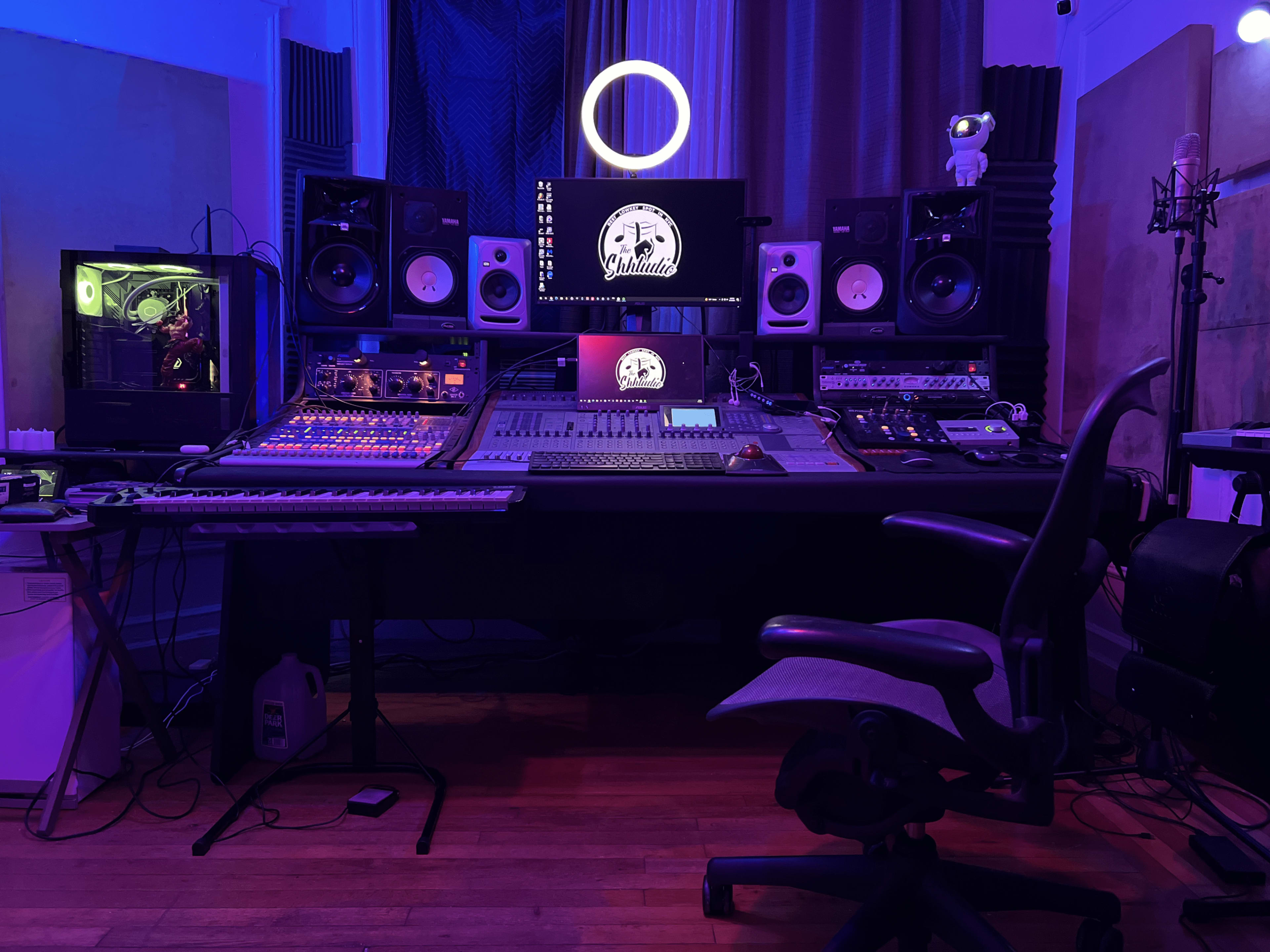 A music studio equipped with a large mixing desk, speakers, multiple audio devices, a computer monitor, and colorful ambient lighting.