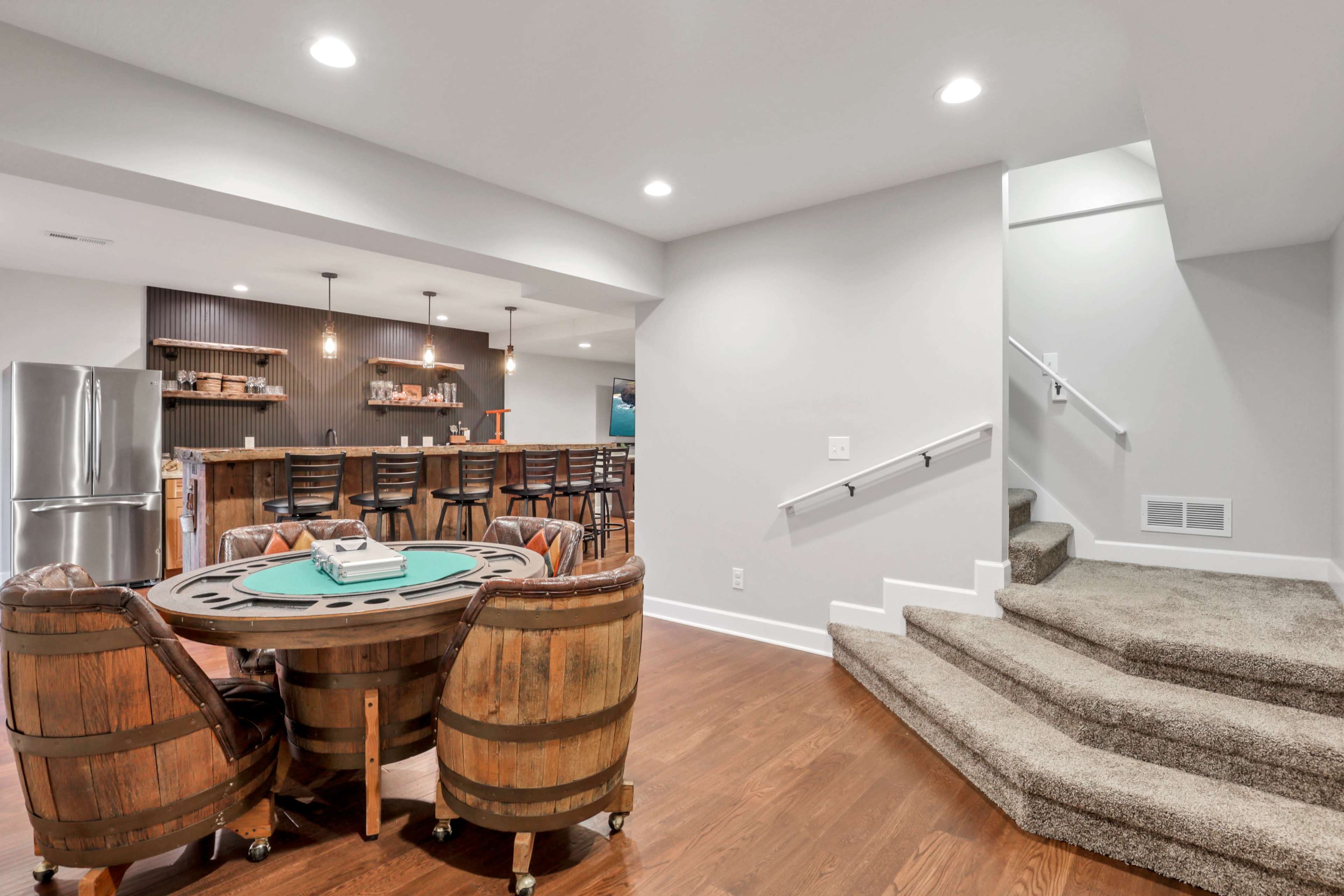 A basement interior featuring a poker table surrounded by wooden barrel chairs, a kitchenette bar in the background, and a staircase leading to an upper level.