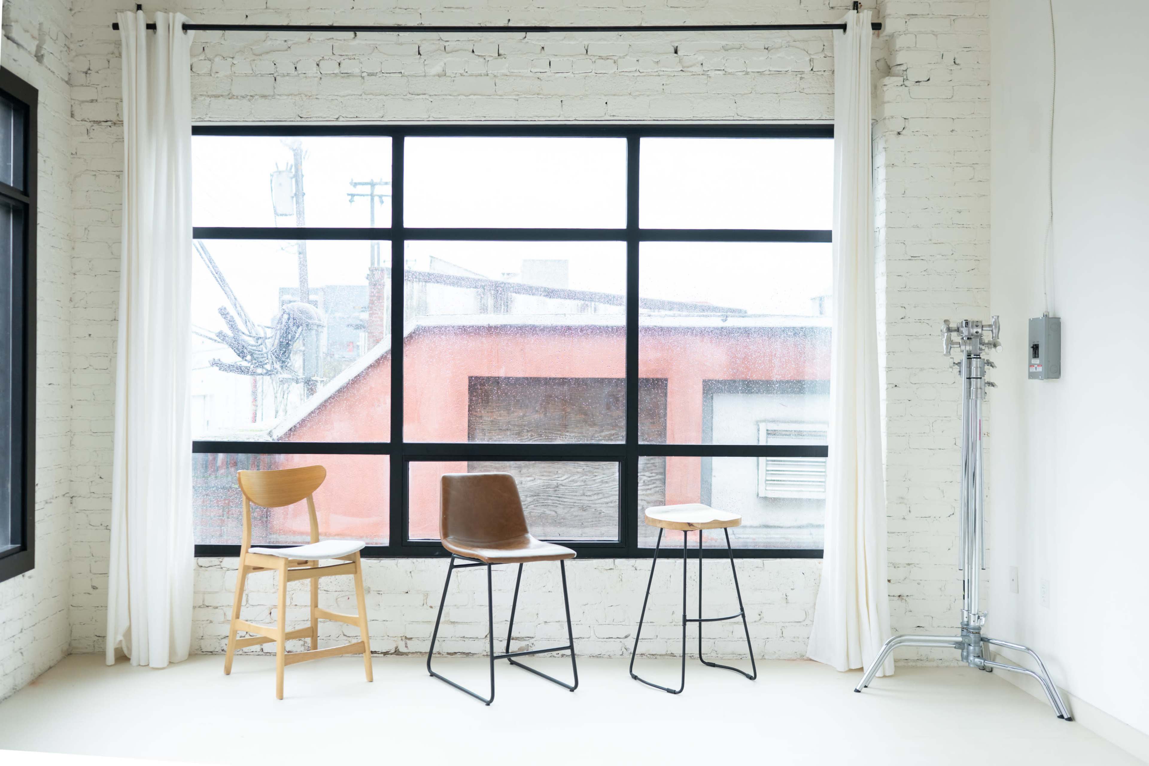 The image shows three chairs of different designs placed against a large window with rain obscuring the view outside, set in a bright, minimalistic room with white brick walls.