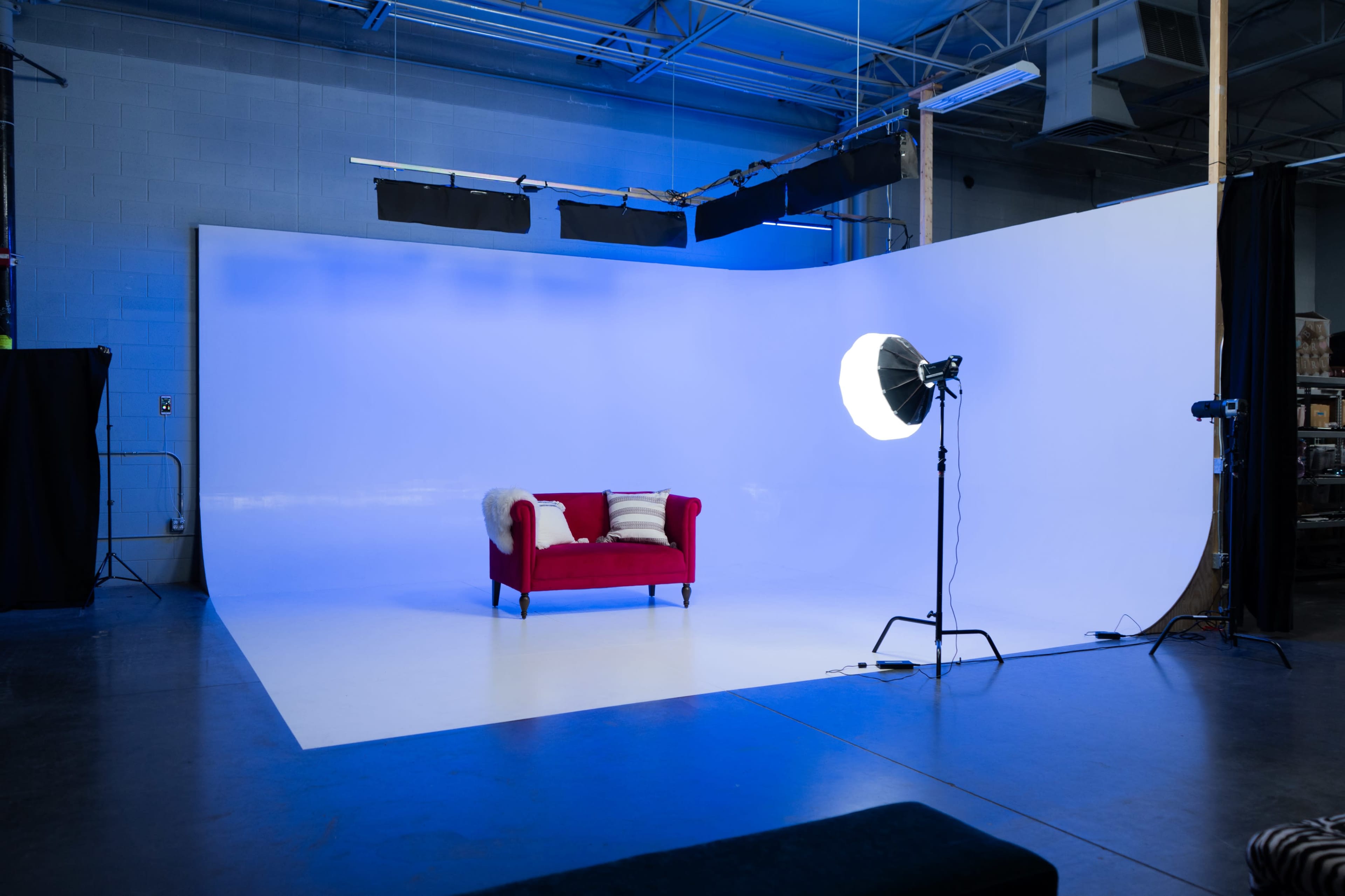 The image shows a photography studio with a red couch in front of a large white backdrop and a lighting setup.