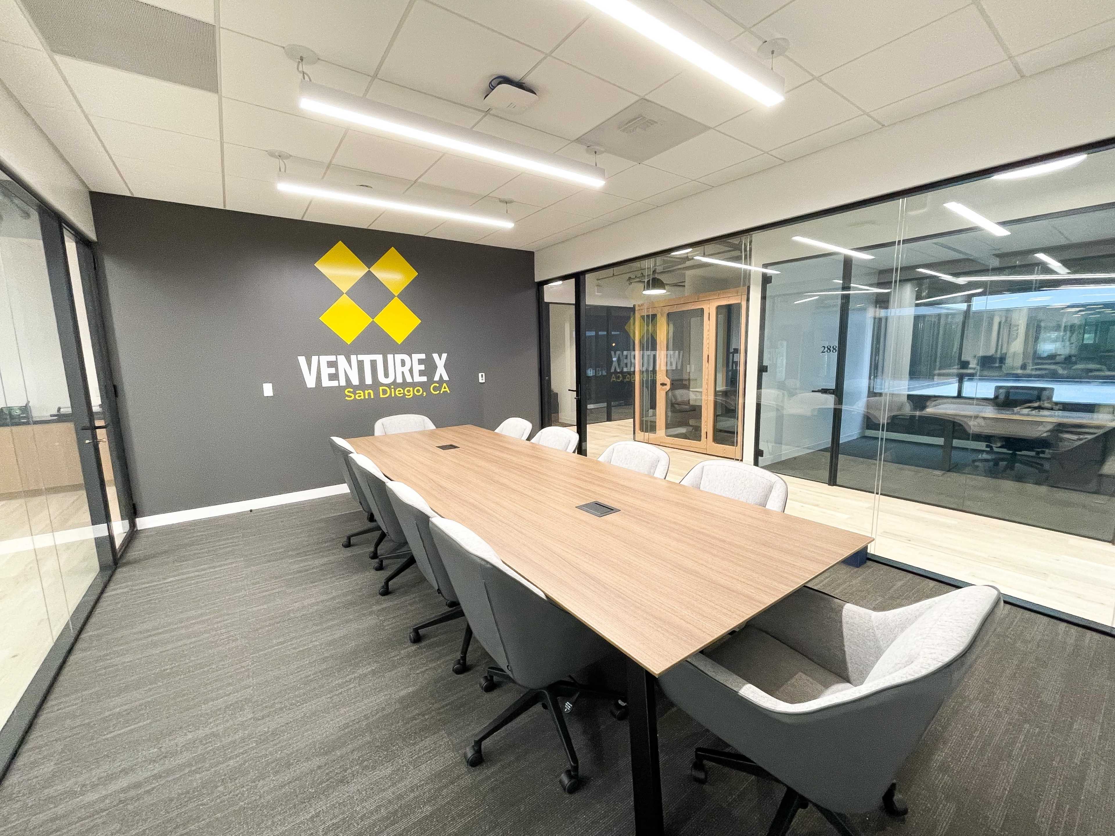 A modern conference room with a long wooden table surrounded by chairs, featuring glass walls and a logo on the wall.