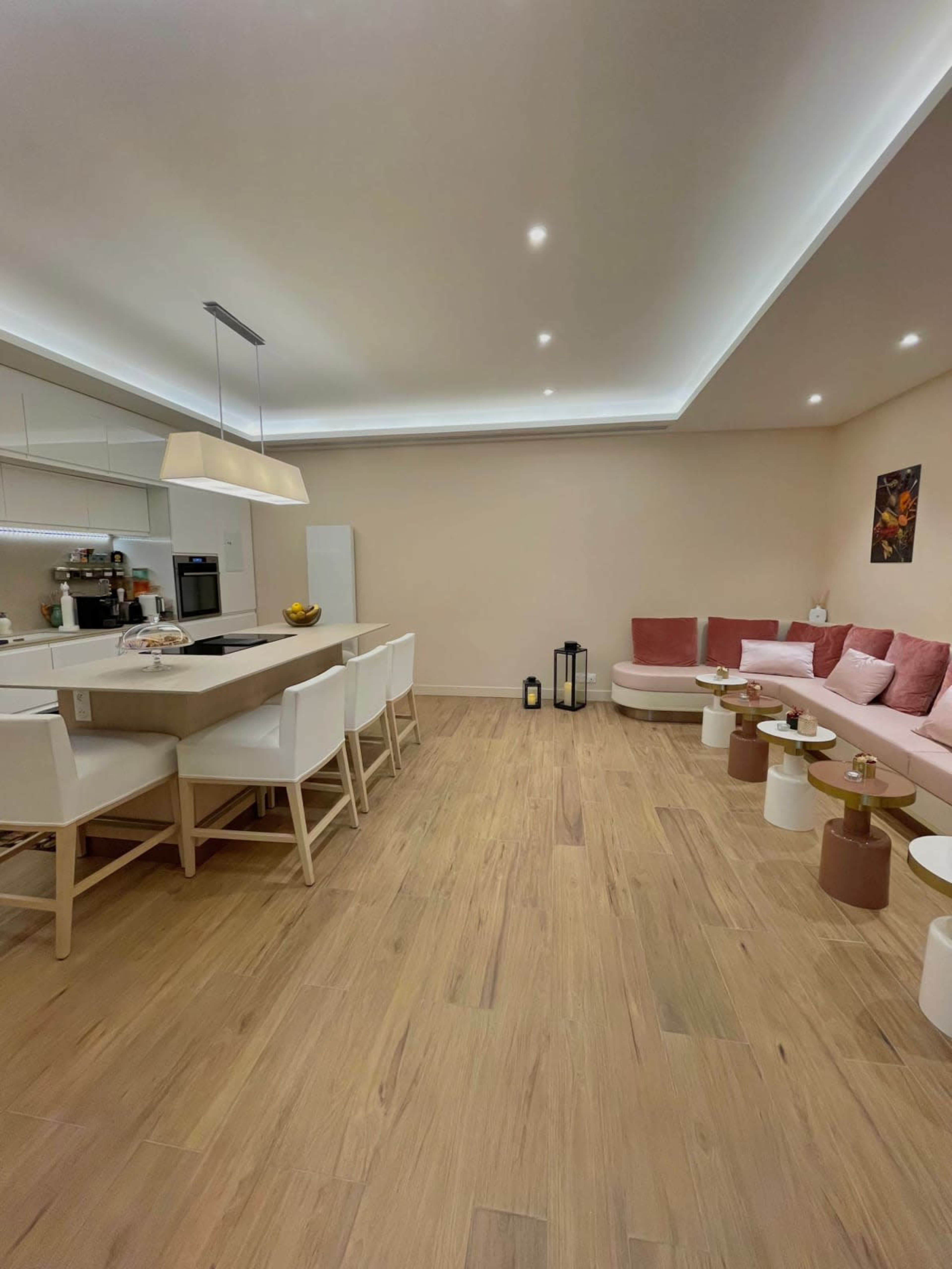 A modern kitchen and living area with a light-colored wooden floor, sleek white cabinetry, a central island, and a seating area featuring pink cushions.