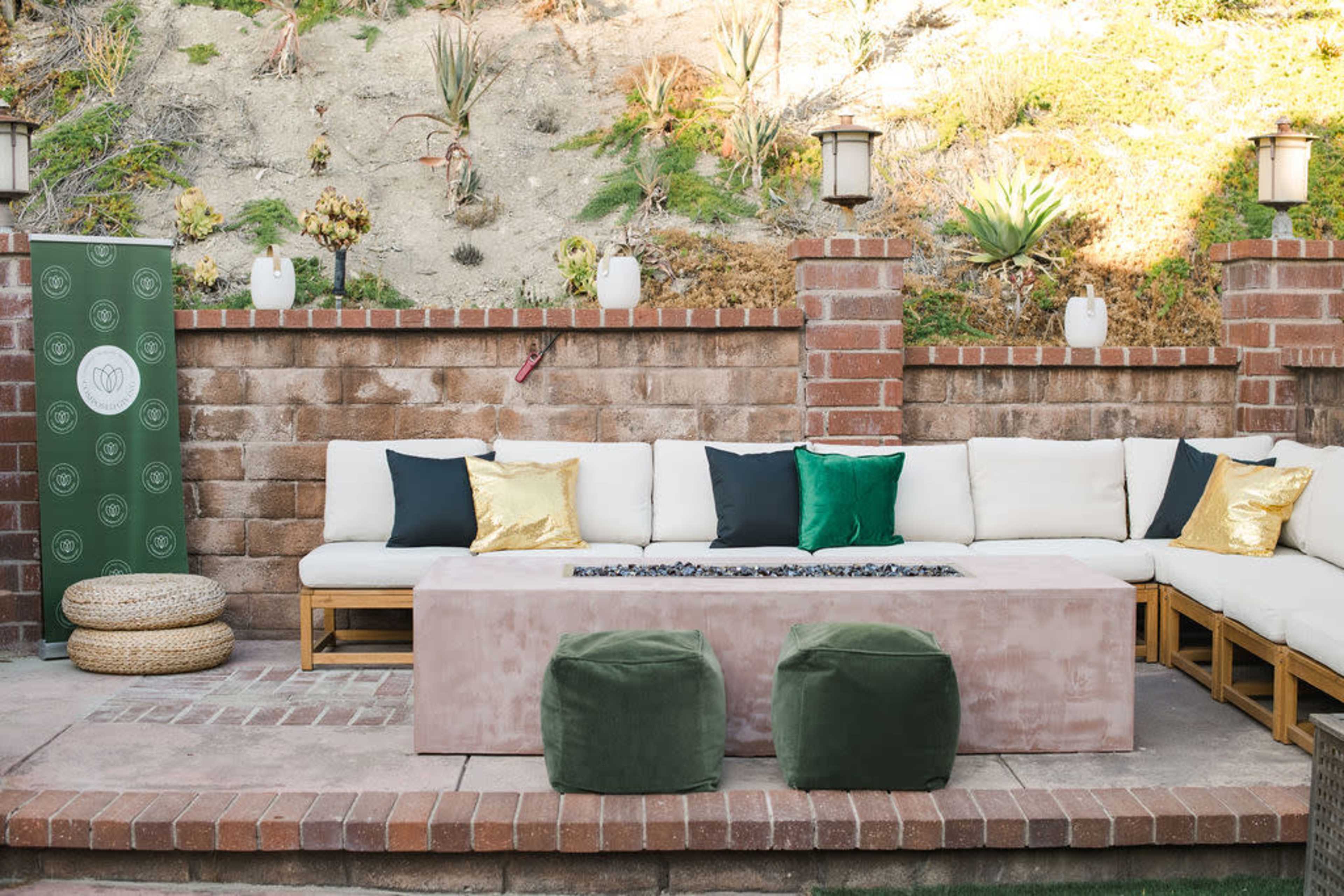 The image shows an outdoor seating area with a long white couch adorned with various colored pillows and a central fire pit surrounded by a stone wall.