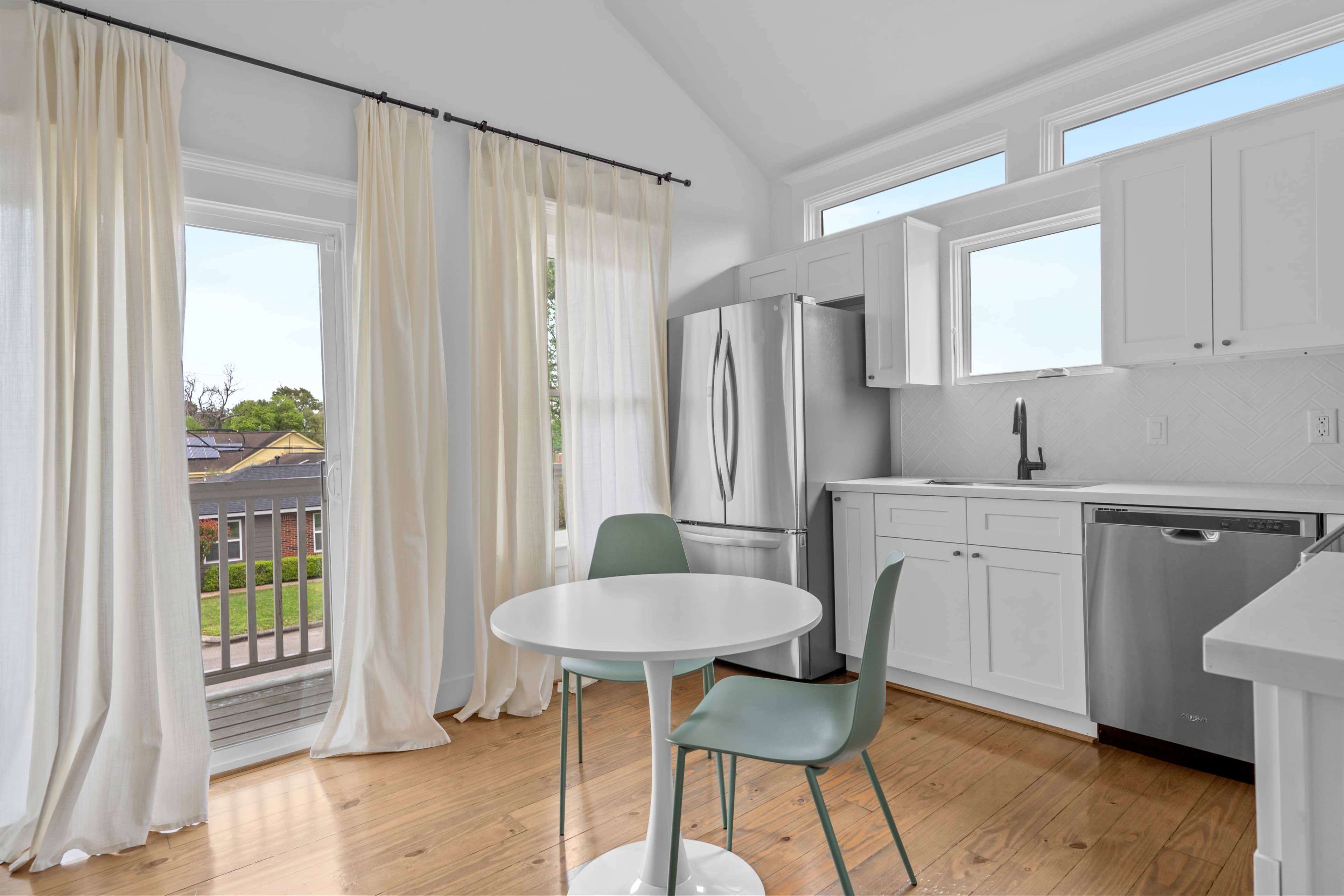 A bright kitchen features a round table with two chairs, a stainless steel refrigerator, and large windows allowing natural light.