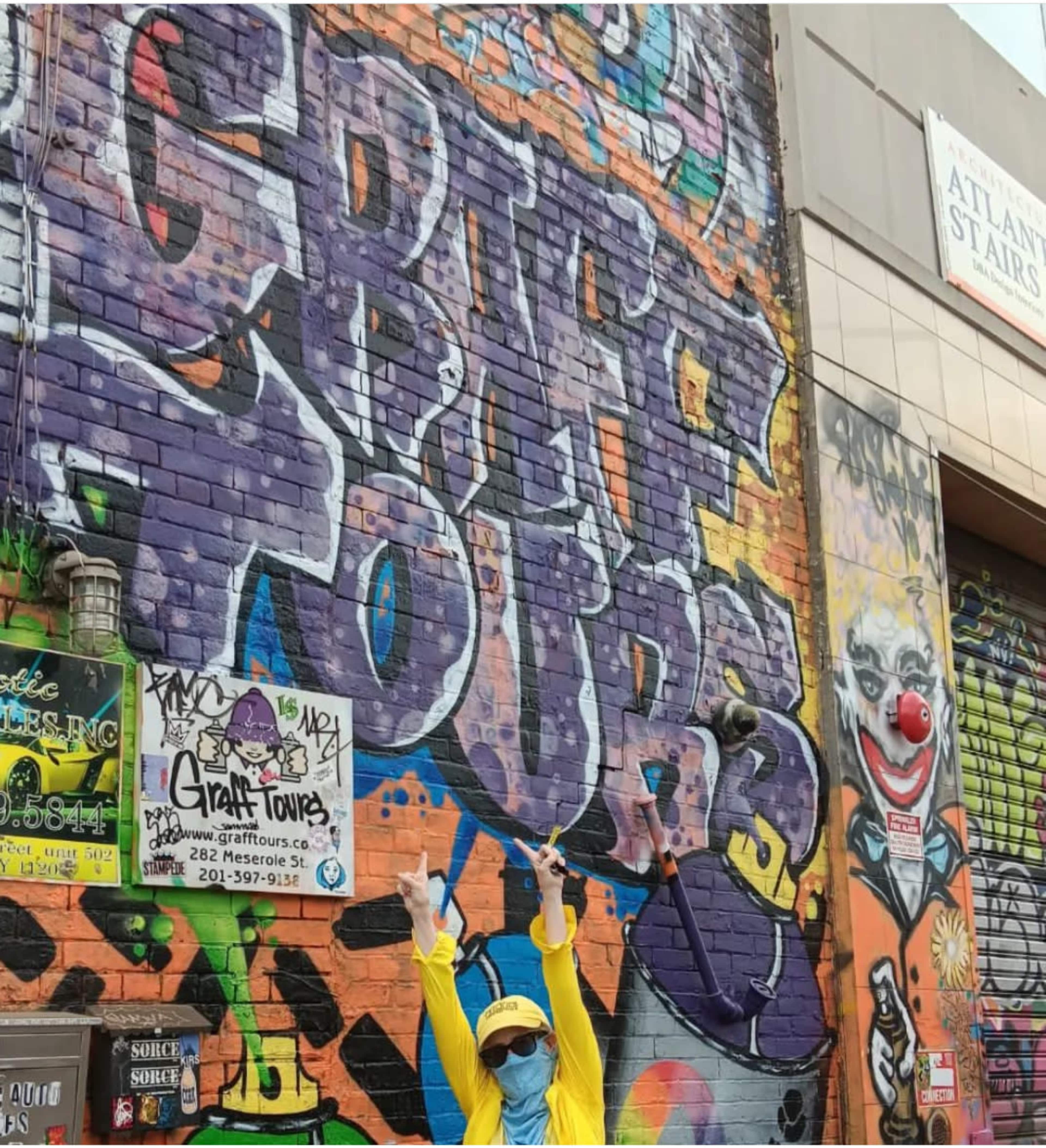 A person in a yellow outfit poses with peace signs in front of a brightly colored graffiti wall that features the words "Graffiti Tours."