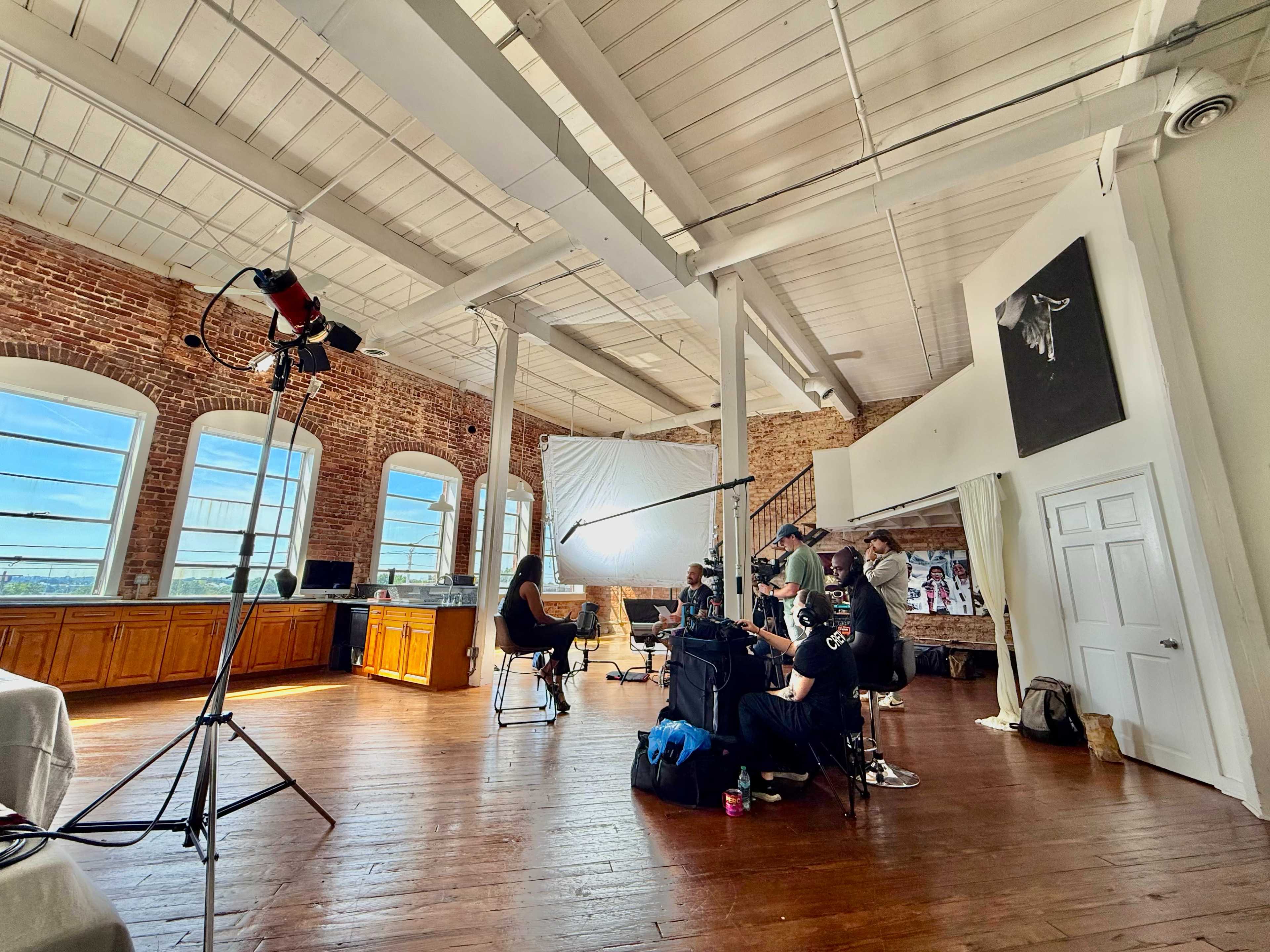 A film crew sets up for a shoot in a spacious room with large windows, brick walls, and wooden floors.