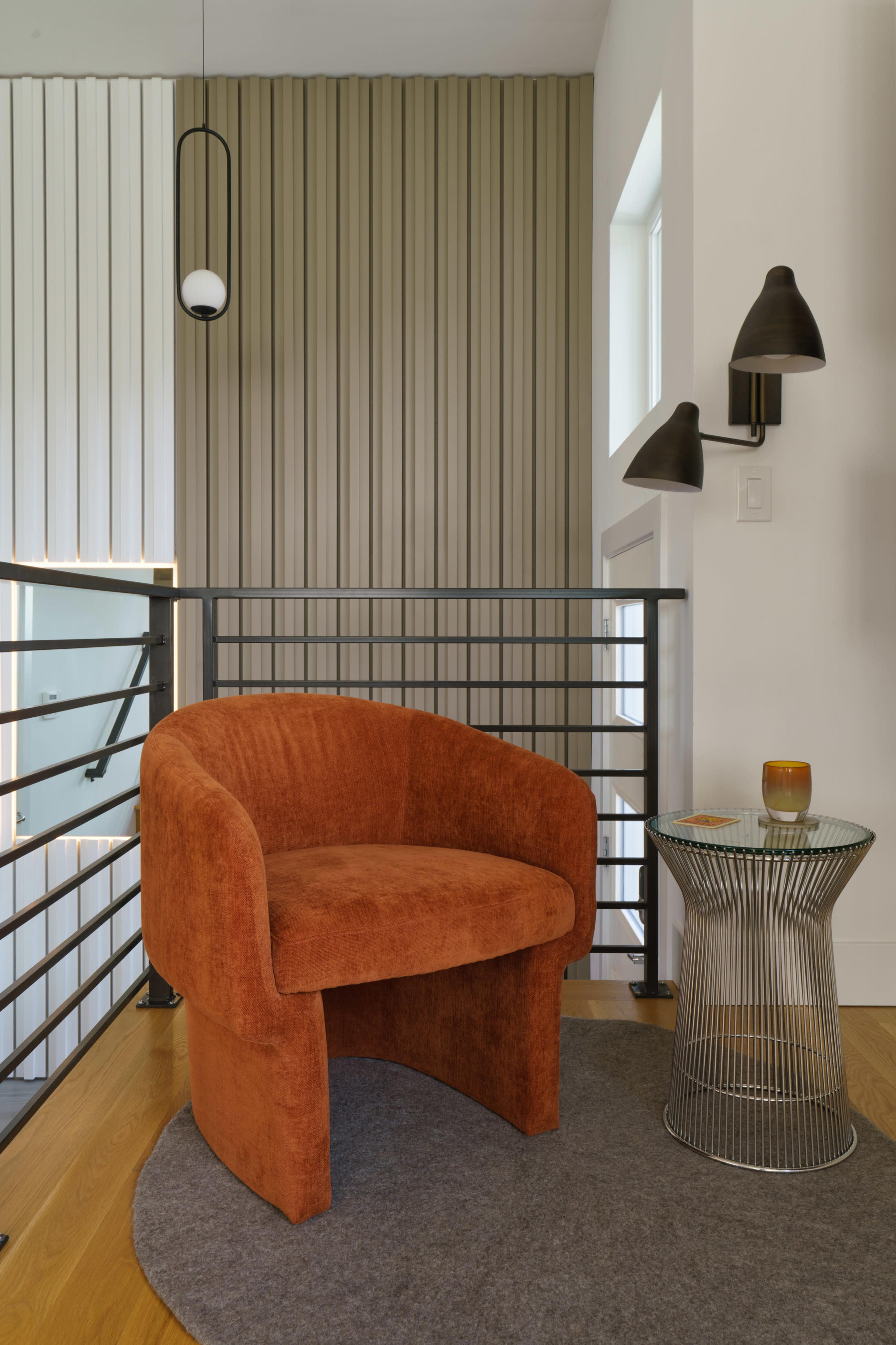 A round orange chair is positioned next to a wire side table on a gray rug in a well-lit interior space with vertical striped walls.