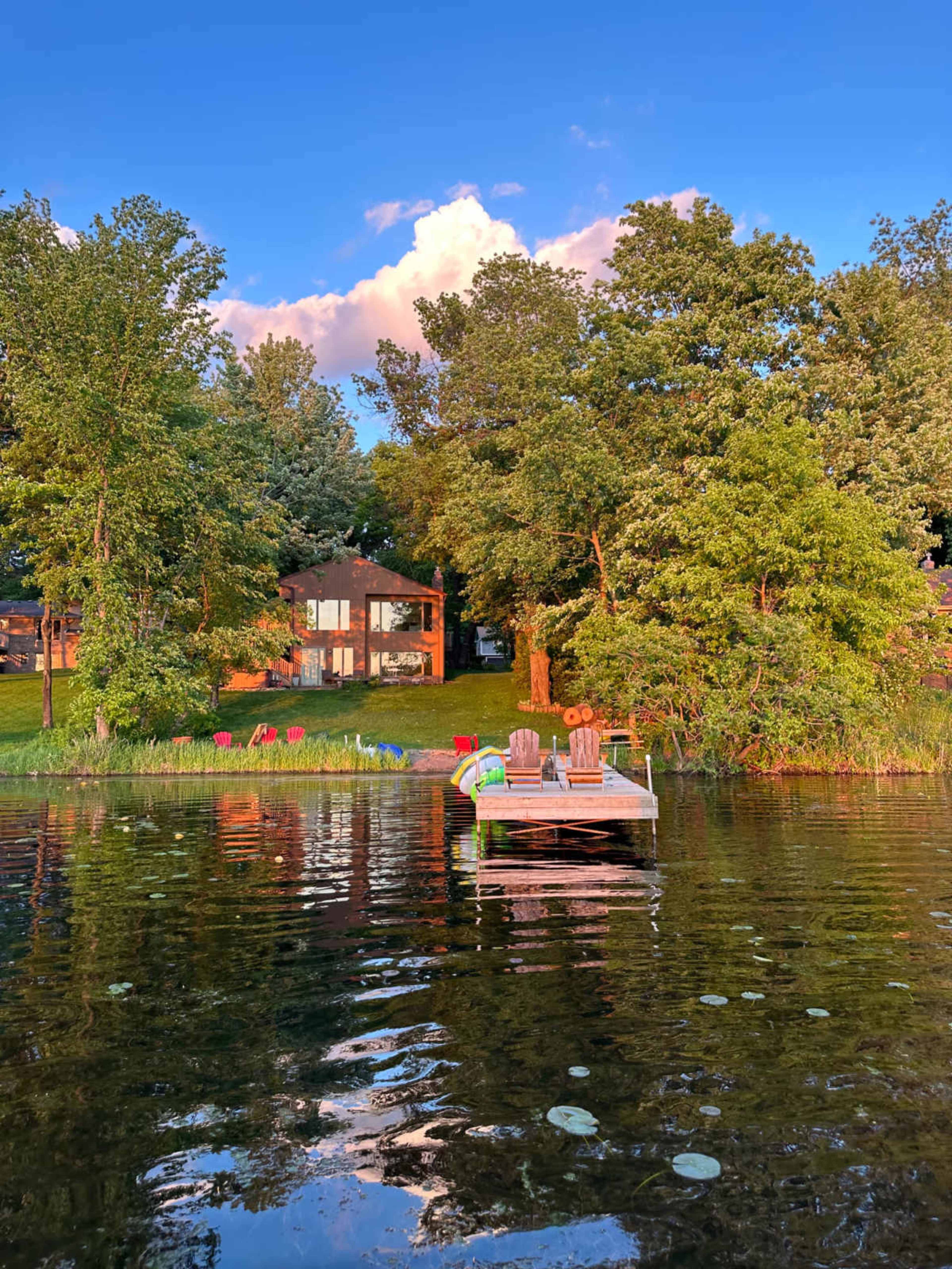 A wooden dock extends into a lake, with a house and green trees reflecting in the water under a blue sky.