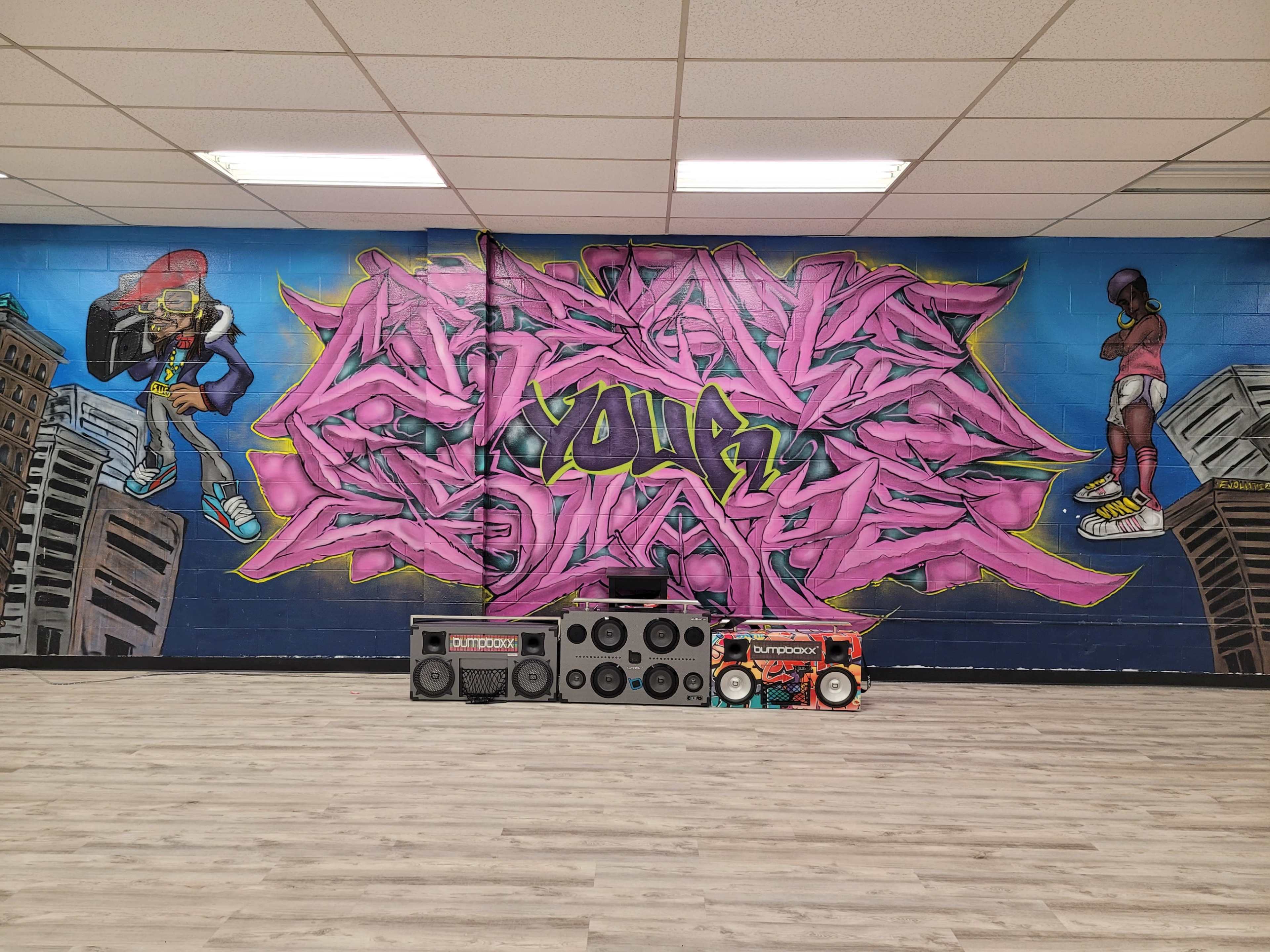 A vibrant mural featuring graffiti-style text and colorful characters is painted on a wall, with several boomboxes arranged in front on a wooden floor.