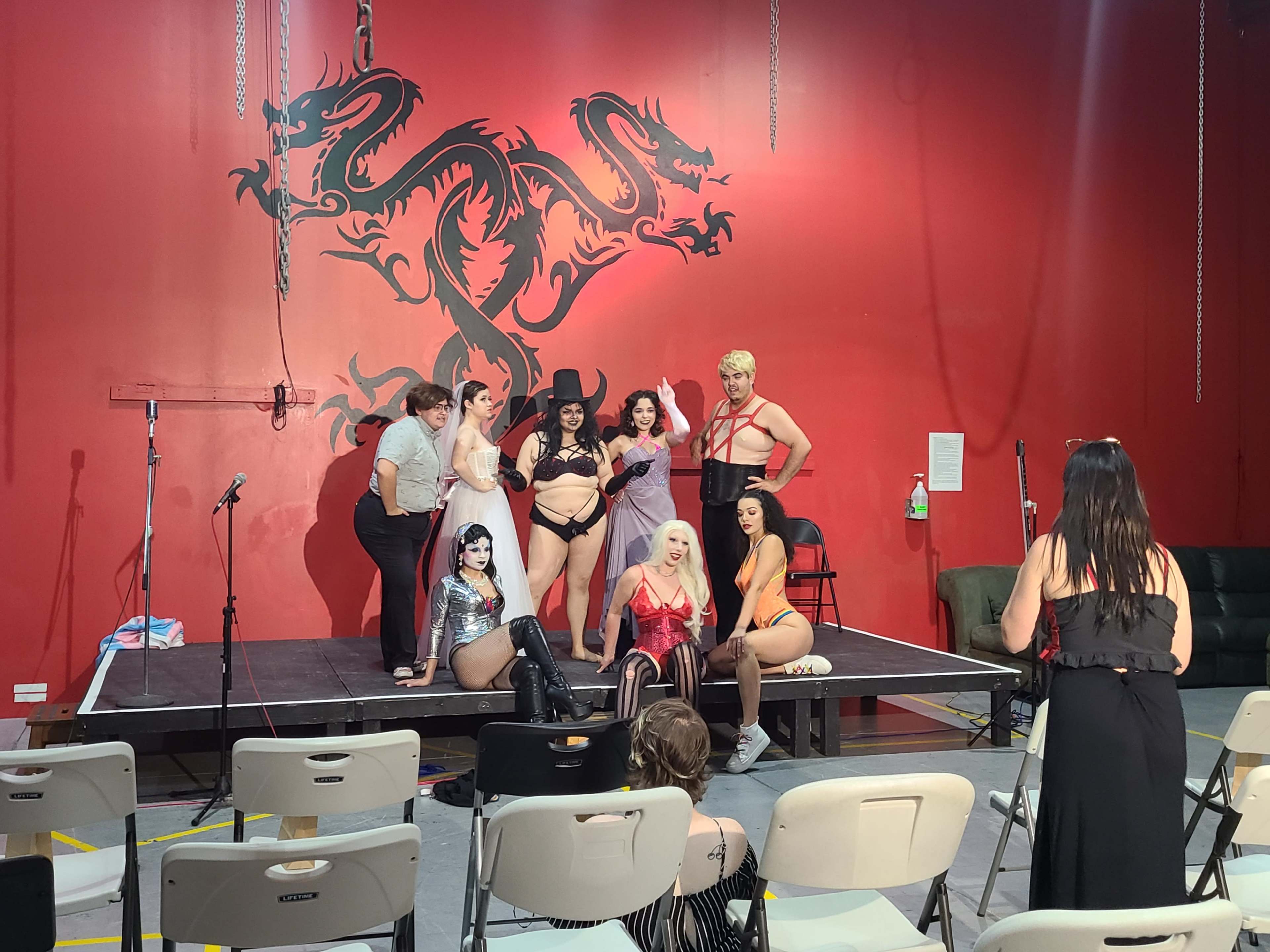 A group of performers poses on a small stage in front of a red wall adorned with a dragon mural.