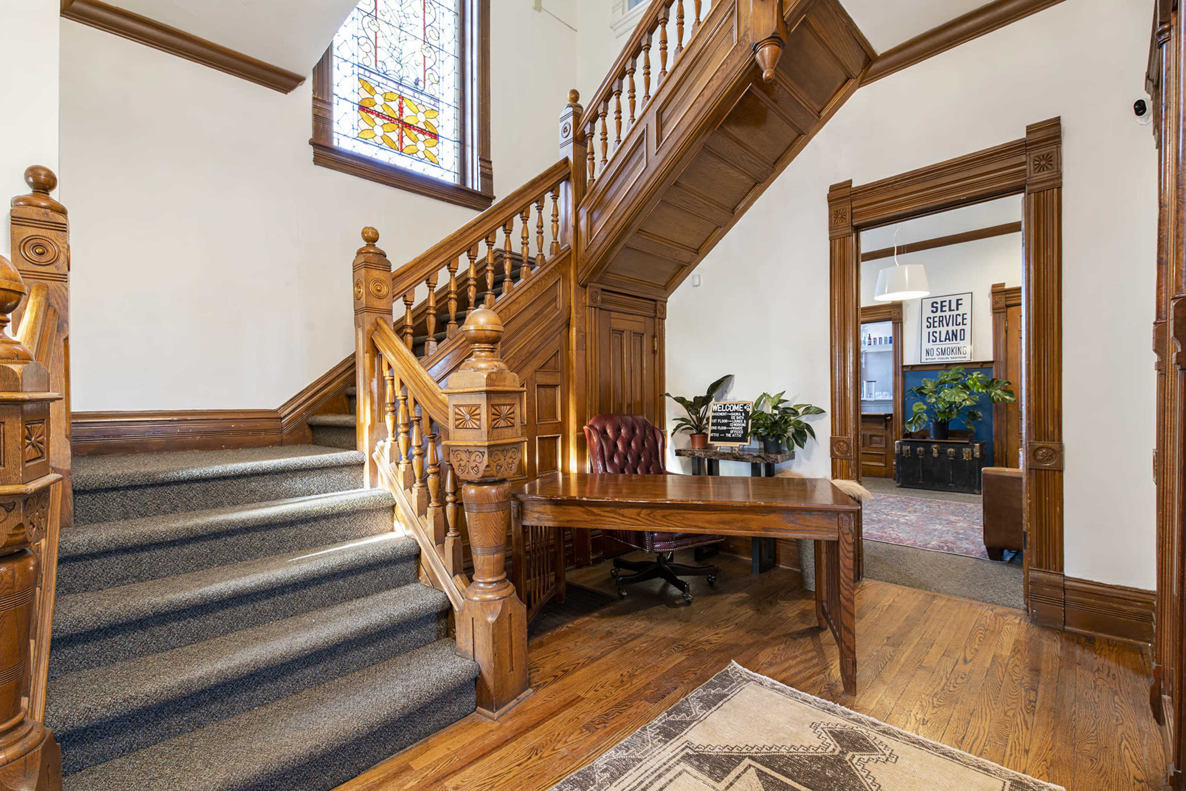 The image shows a wooden staircase with intricate balustrades leading up, next to a reception area with a desk and potted plants.