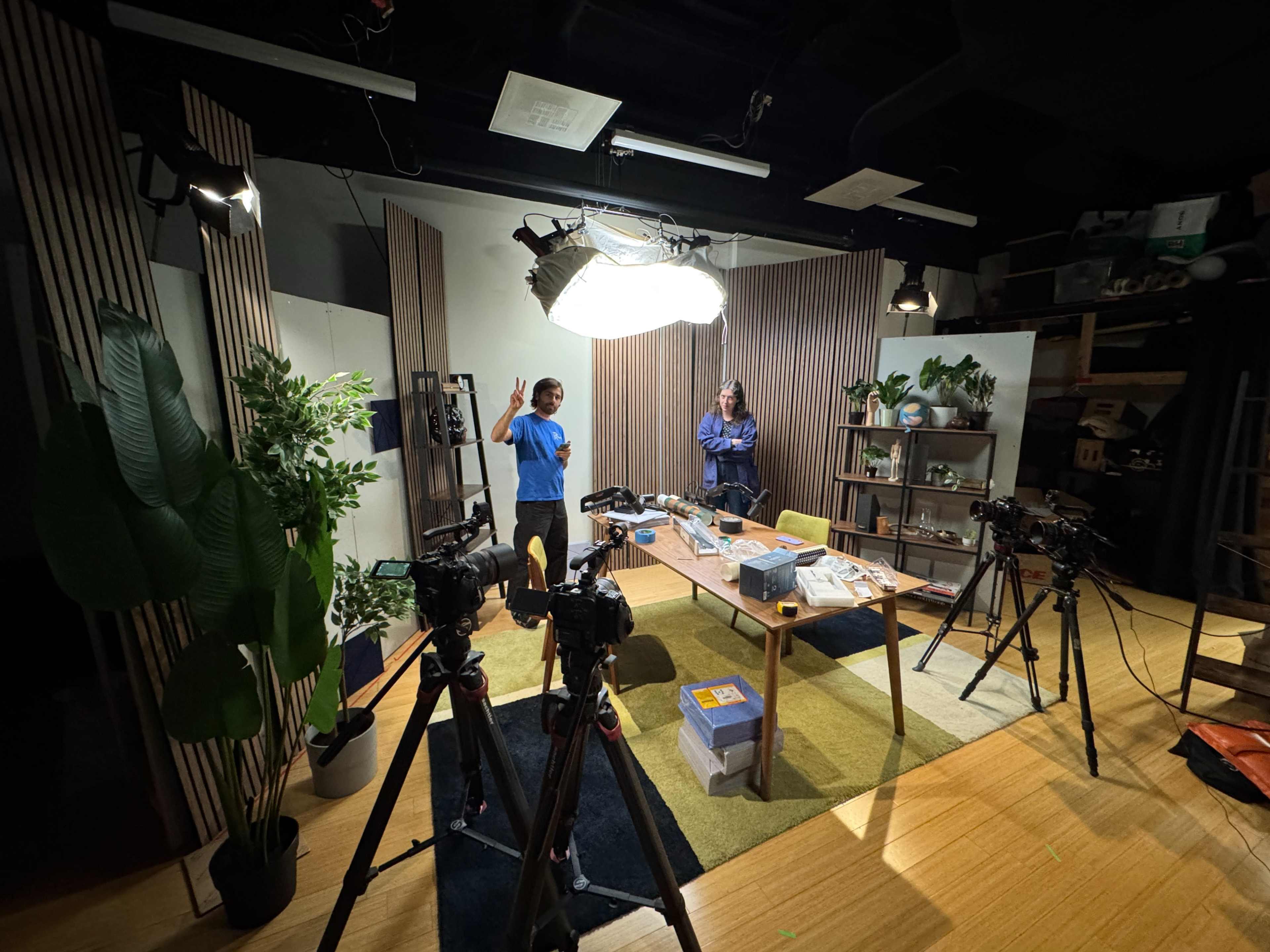 The image shows a brightly lit studio setup with two people, a table covered in various items, and three cameras positioned around the space.