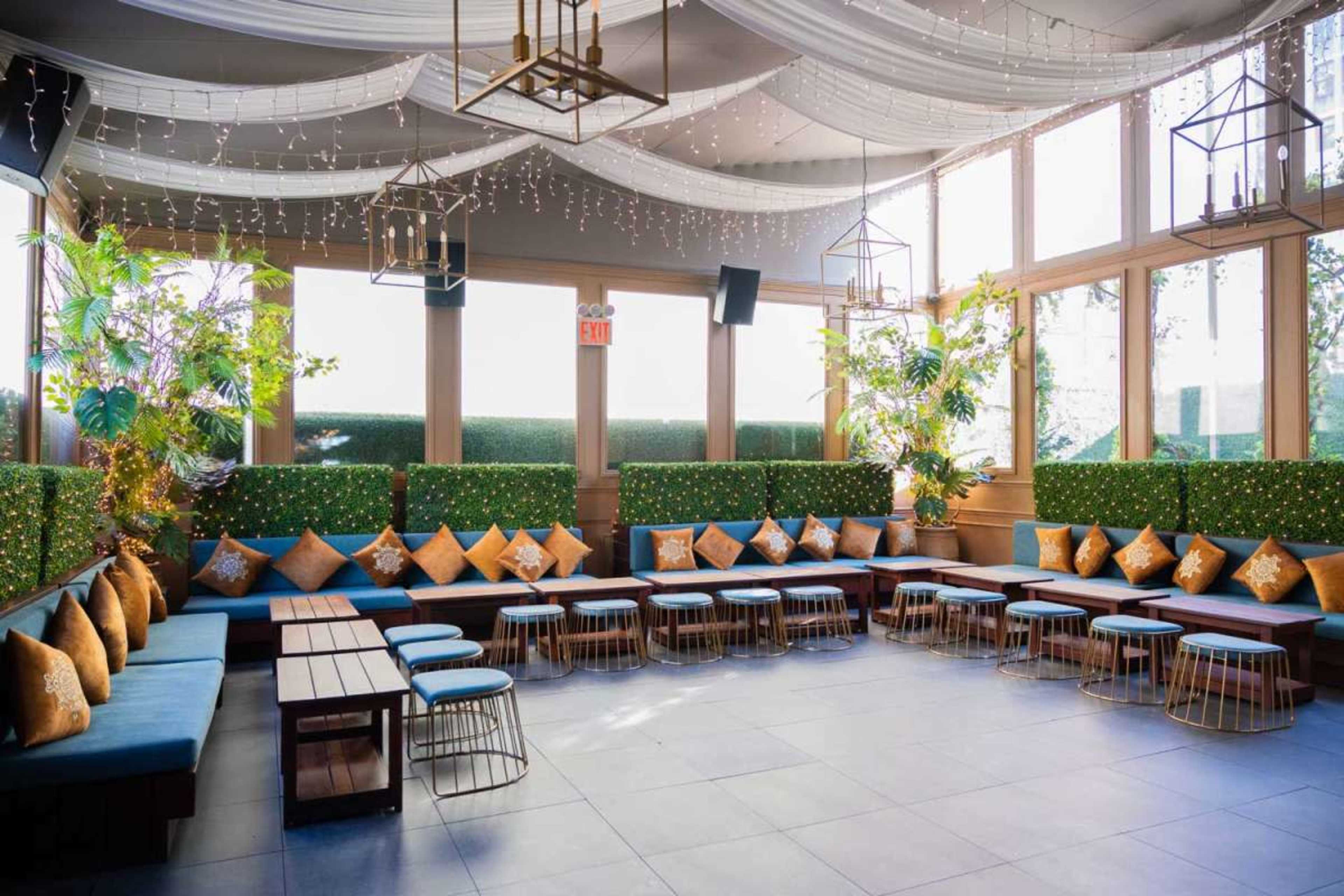 The image shows a modern indoor lounge area with blue seating, wooden tables, and potted plants, illuminated by hanging lights.