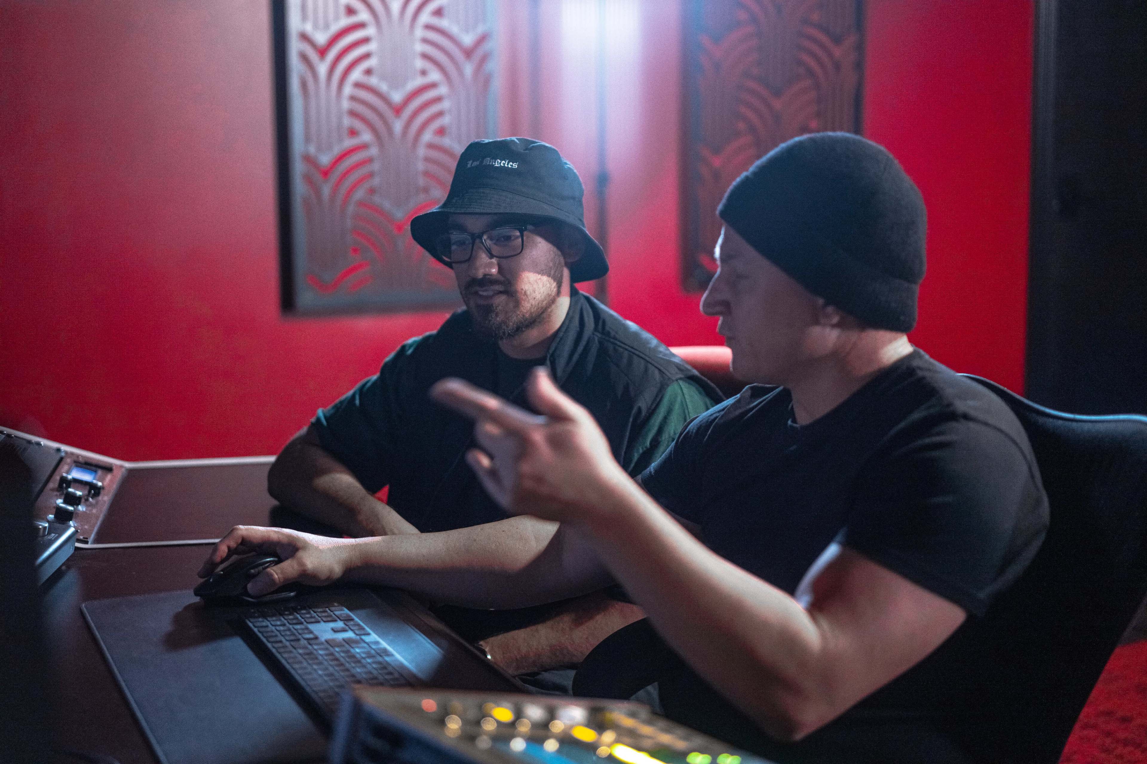 Two men are discussing sound production while sitting at a computer in a dimly lit recording studio.