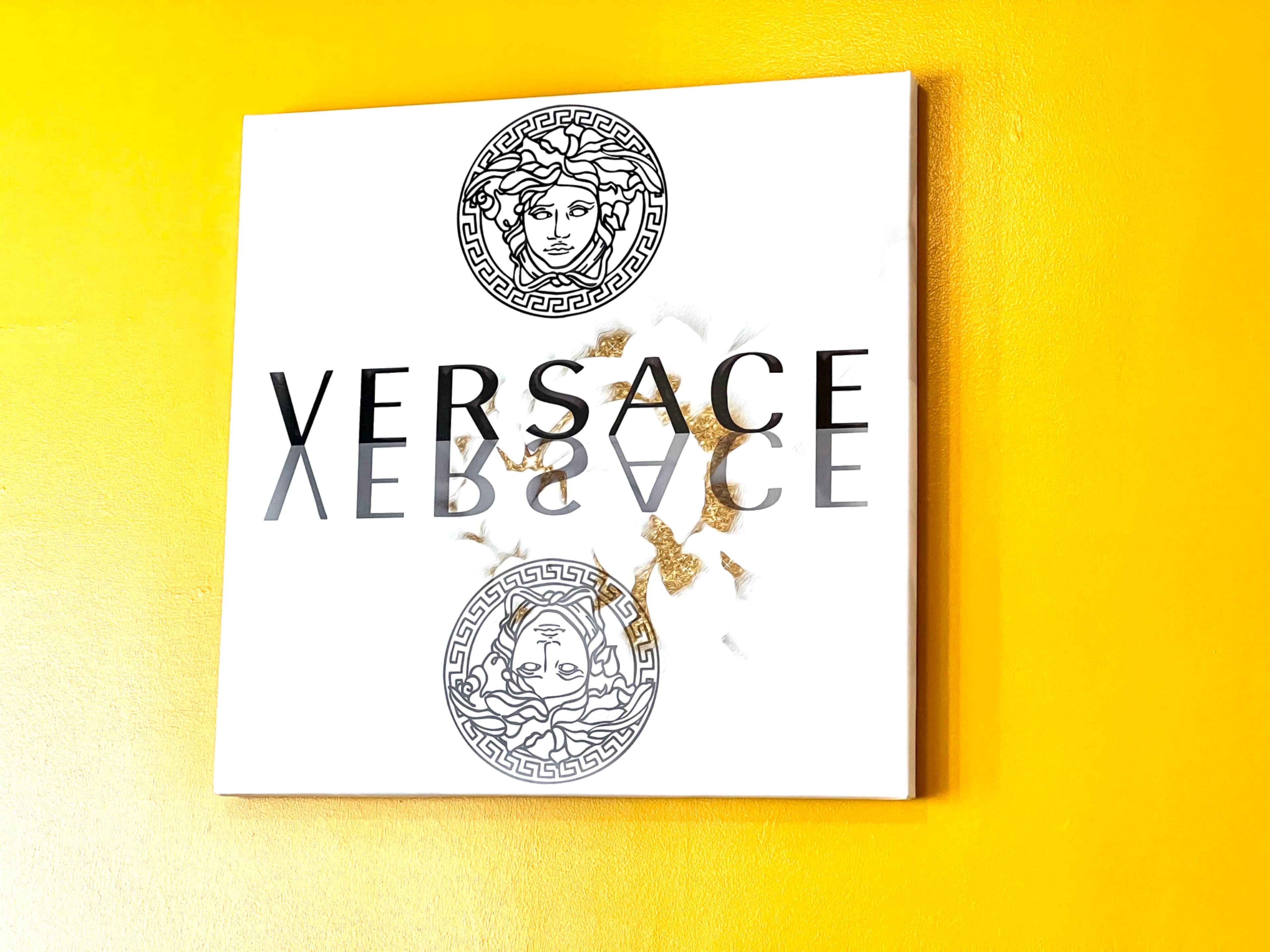 A bold Versace logo on a white background, positioned against a vibrant yellow wall, with some scattered gold flakes.