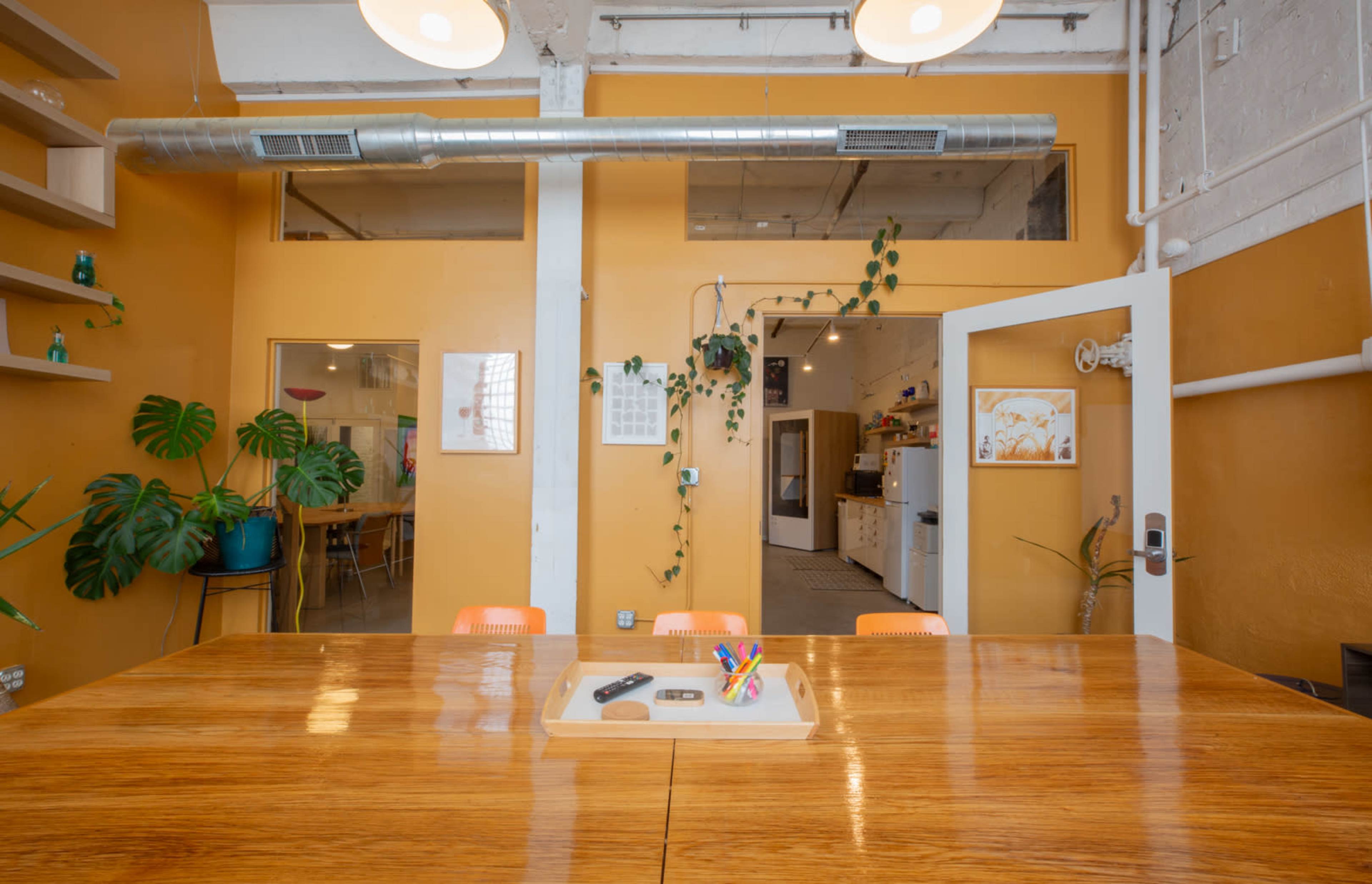 A large wooden table with an assortment of pens sits in a brightly colored room featuring a kitchenette and plants along the walls.