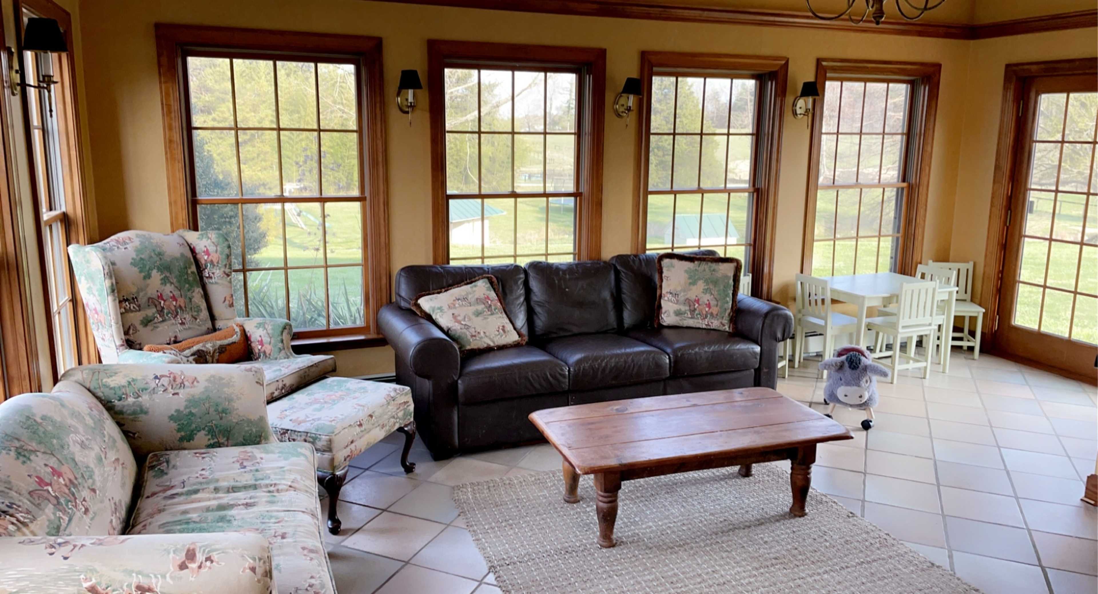 The image shows a sunroom furnished with a dark leather sofa, floral armchairs, a wooden coffee table, and a small dining set, all surrounded by large windows that provide a view of the outdoors.