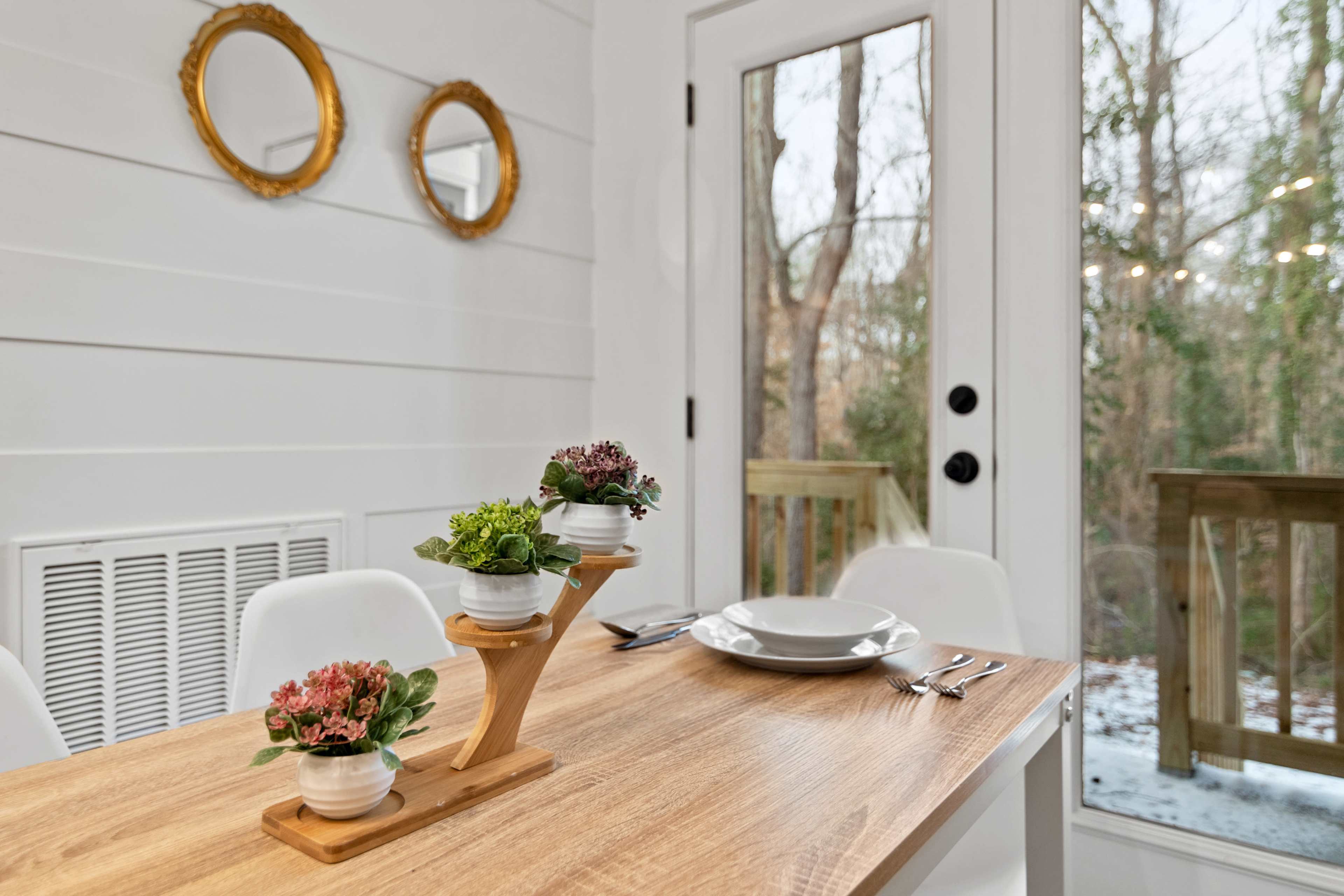 A dining table set with plates and utensils is positioned near a sliding glass door, adorned with potted plants on a wooden stand and framed mirrors on the wall.