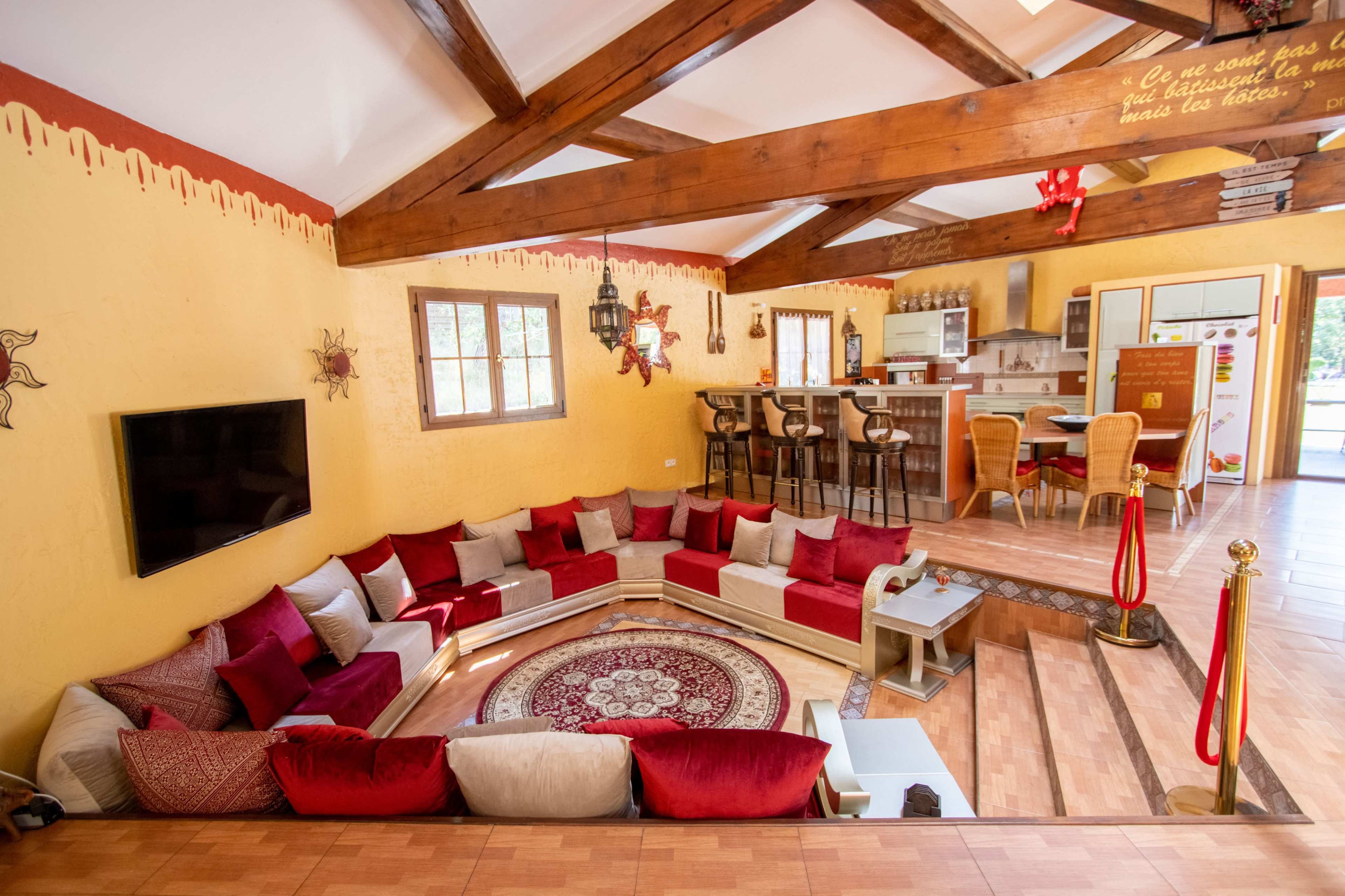 The image shows a cozy, sunlit living area with a circular seating arrangement surrounded by decorative cushions, exposed wooden beams, and a kitchen in the background.