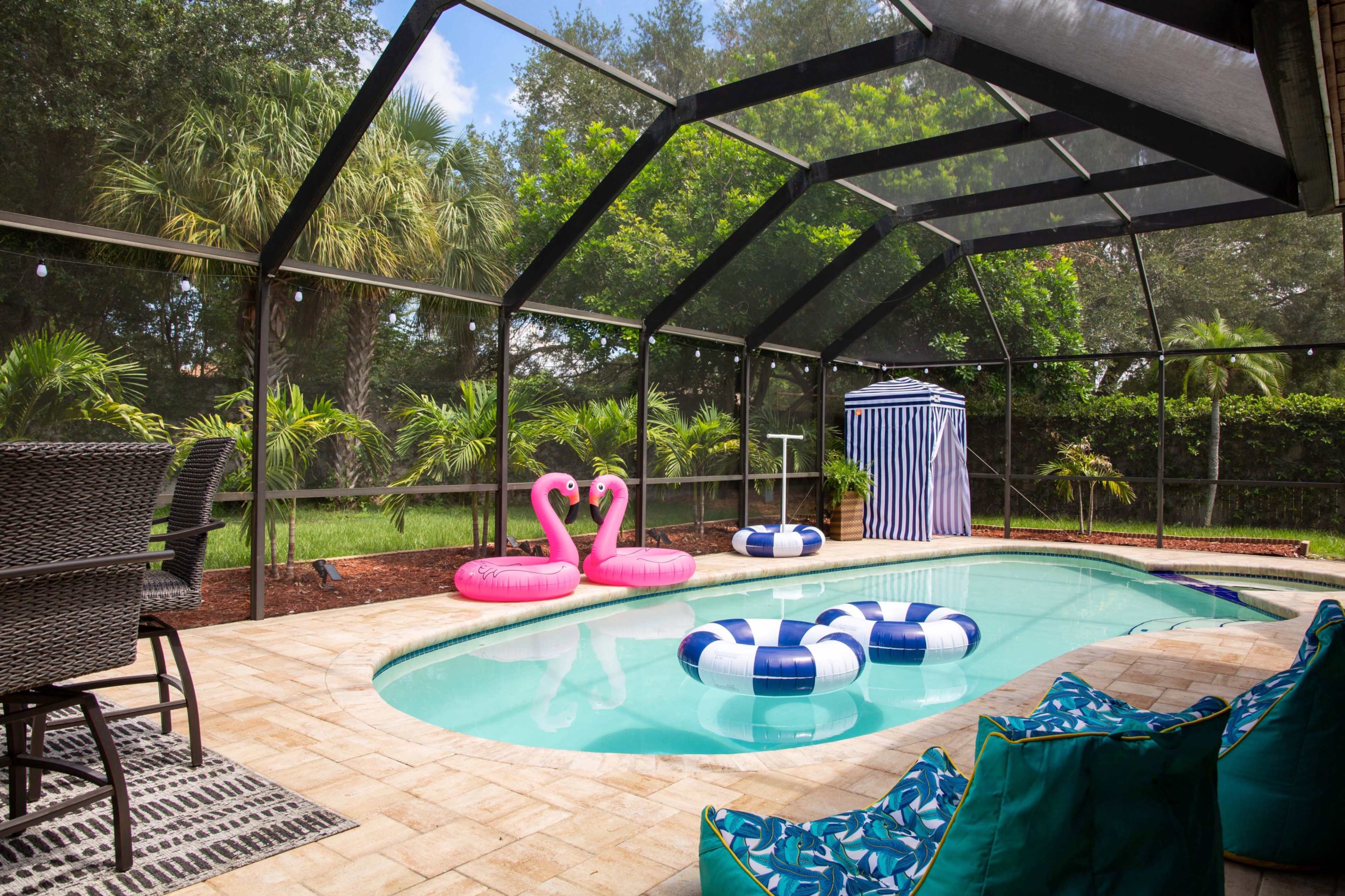 A screened-in pool area with a swimming pool, pink flamingo floaties, blue and white striped float, a changing tent, and outdoor furniture surrounded by greenery.
