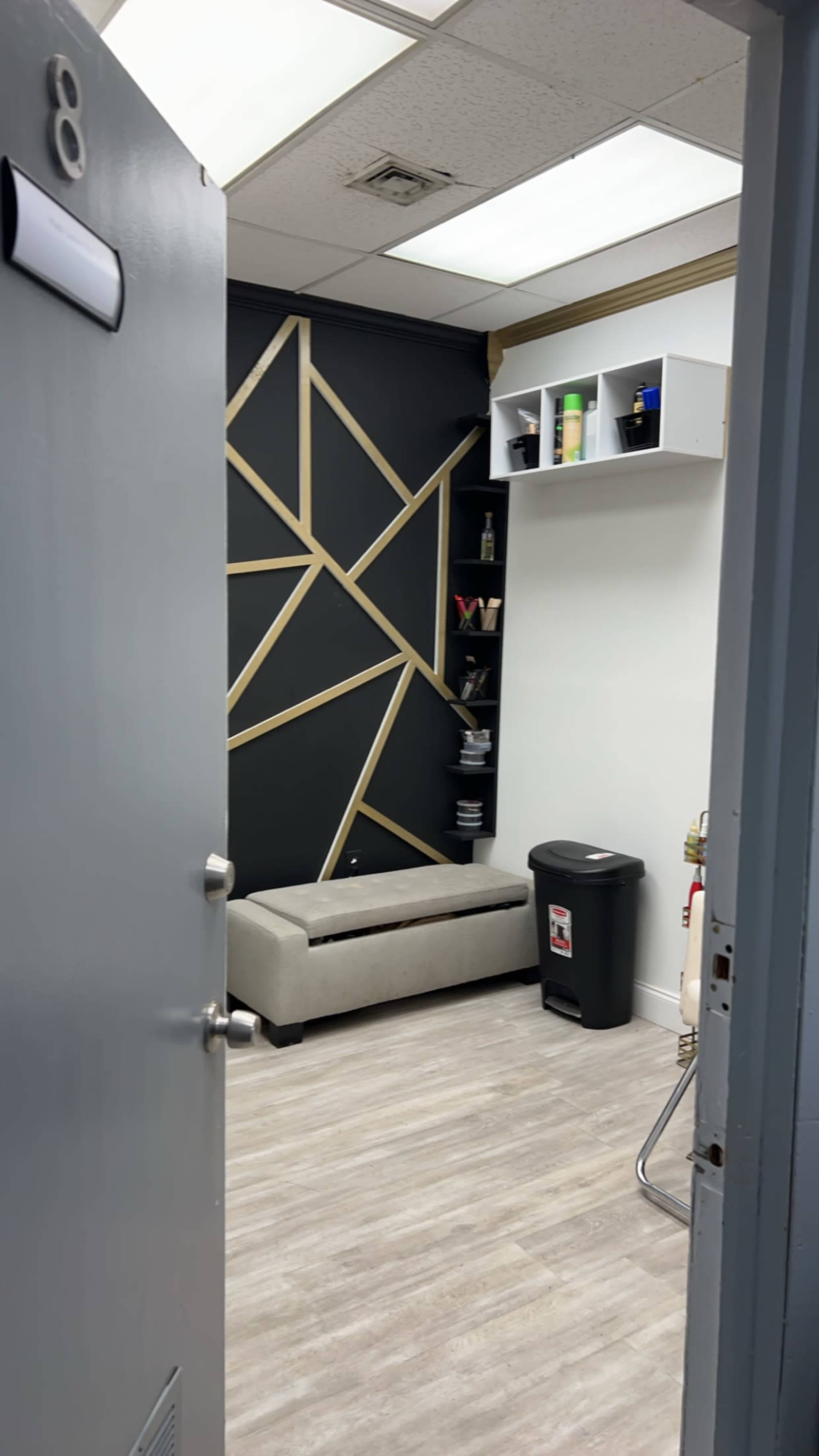 A small waiting area is visible through an open door, featuring a gray bench, a black trash can, and a geometric wall design with shelves displaying various items.
