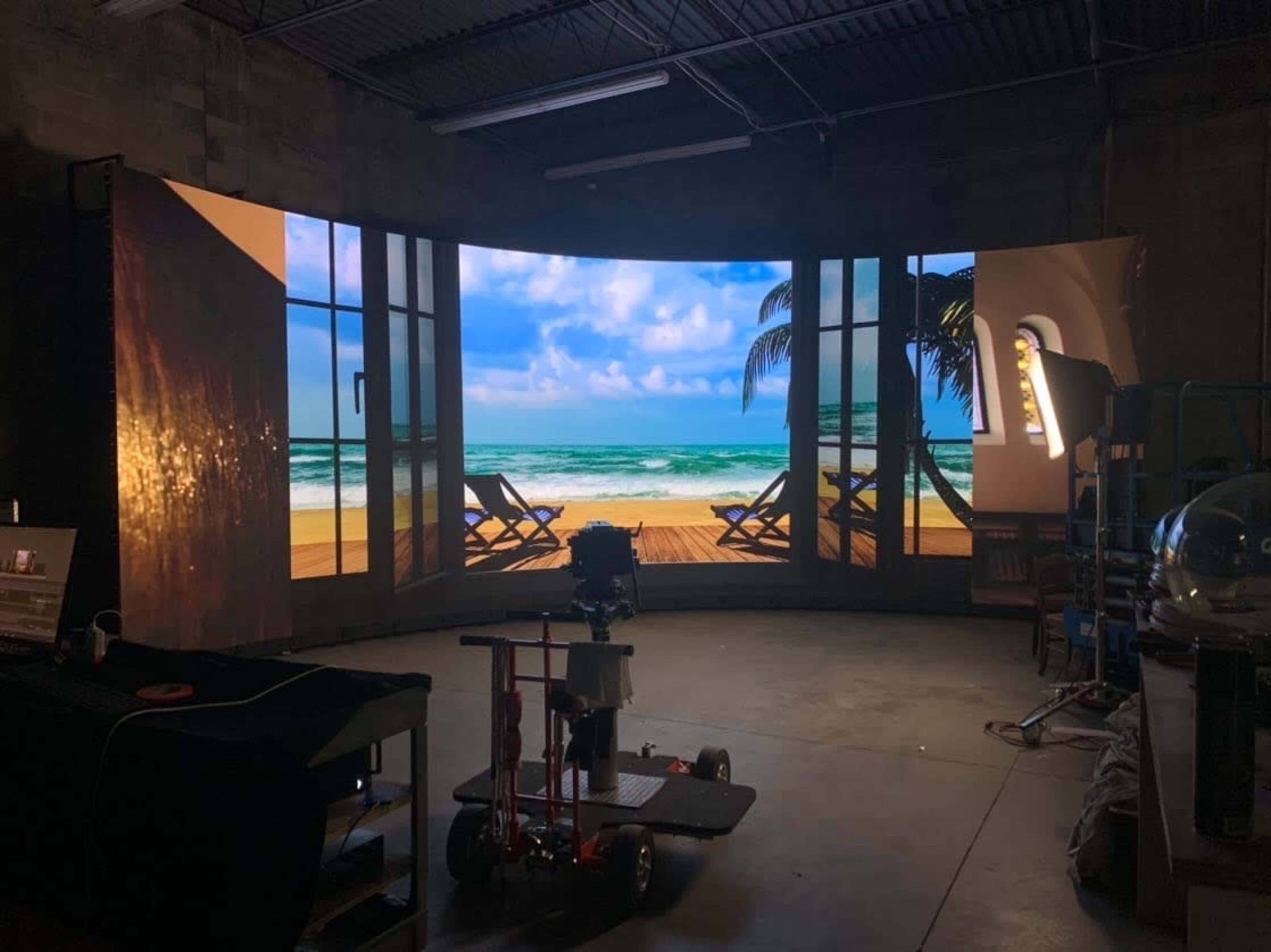 A production studio with a large curved screen displaying a beach scene, including sandy shores and lounge chairs.