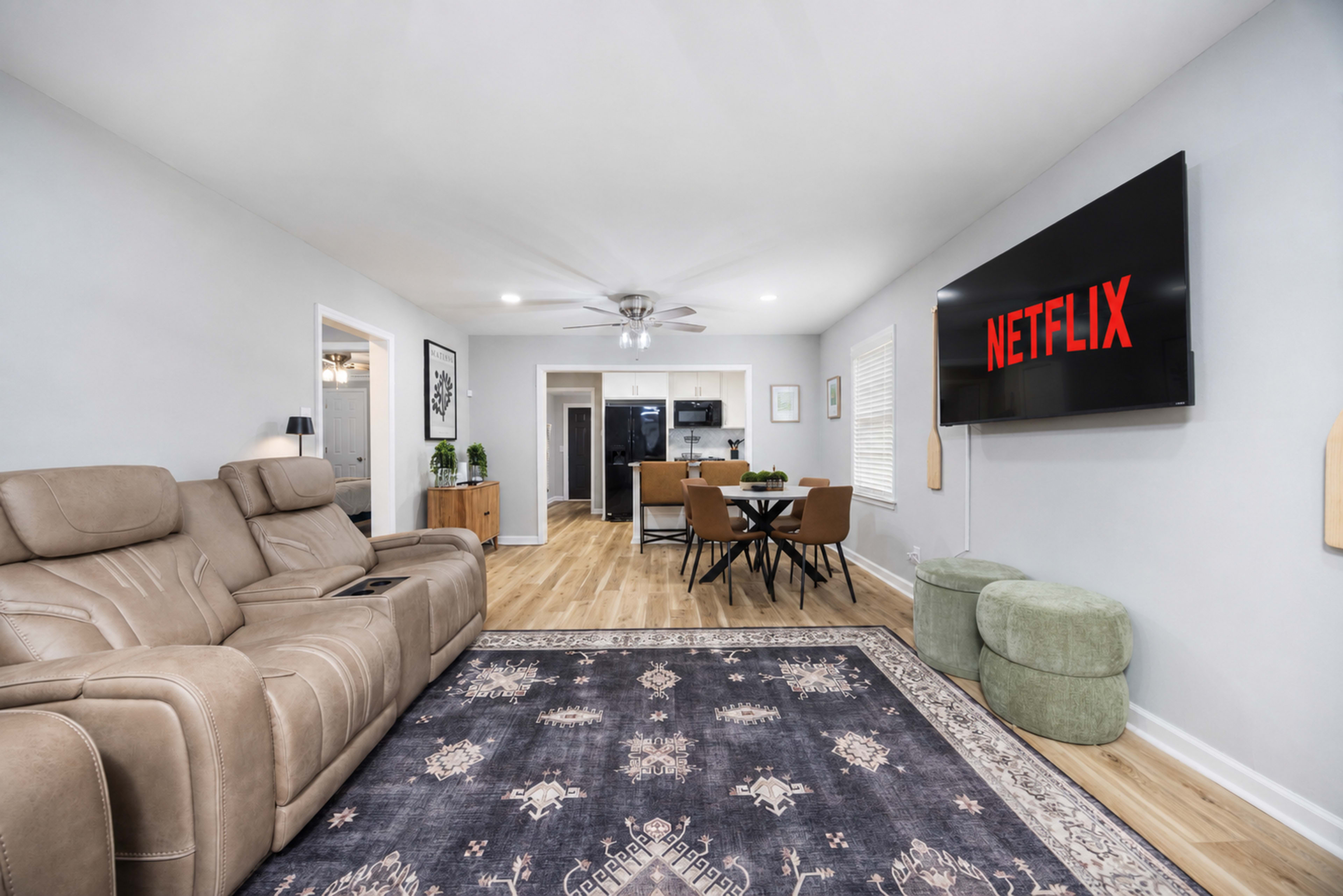 The living room features a large screen displaying the Netflix logo, with a sectional sofa, a dining area in the background, and light wood flooring.