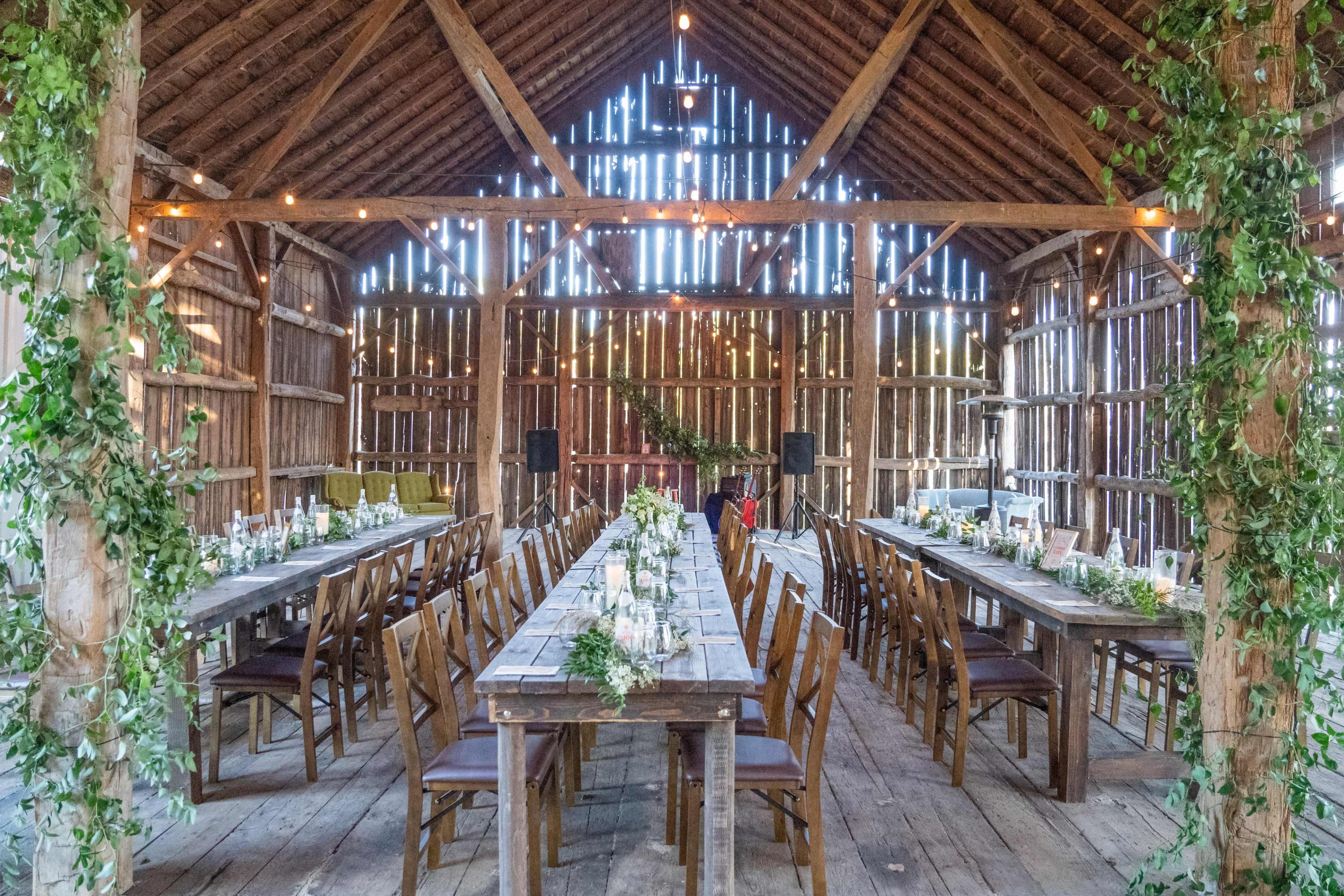 The interior of a barn features two long wooden tables set with candles and floral arrangements, flanked by chairs, under a ceiling adorned with string lights.