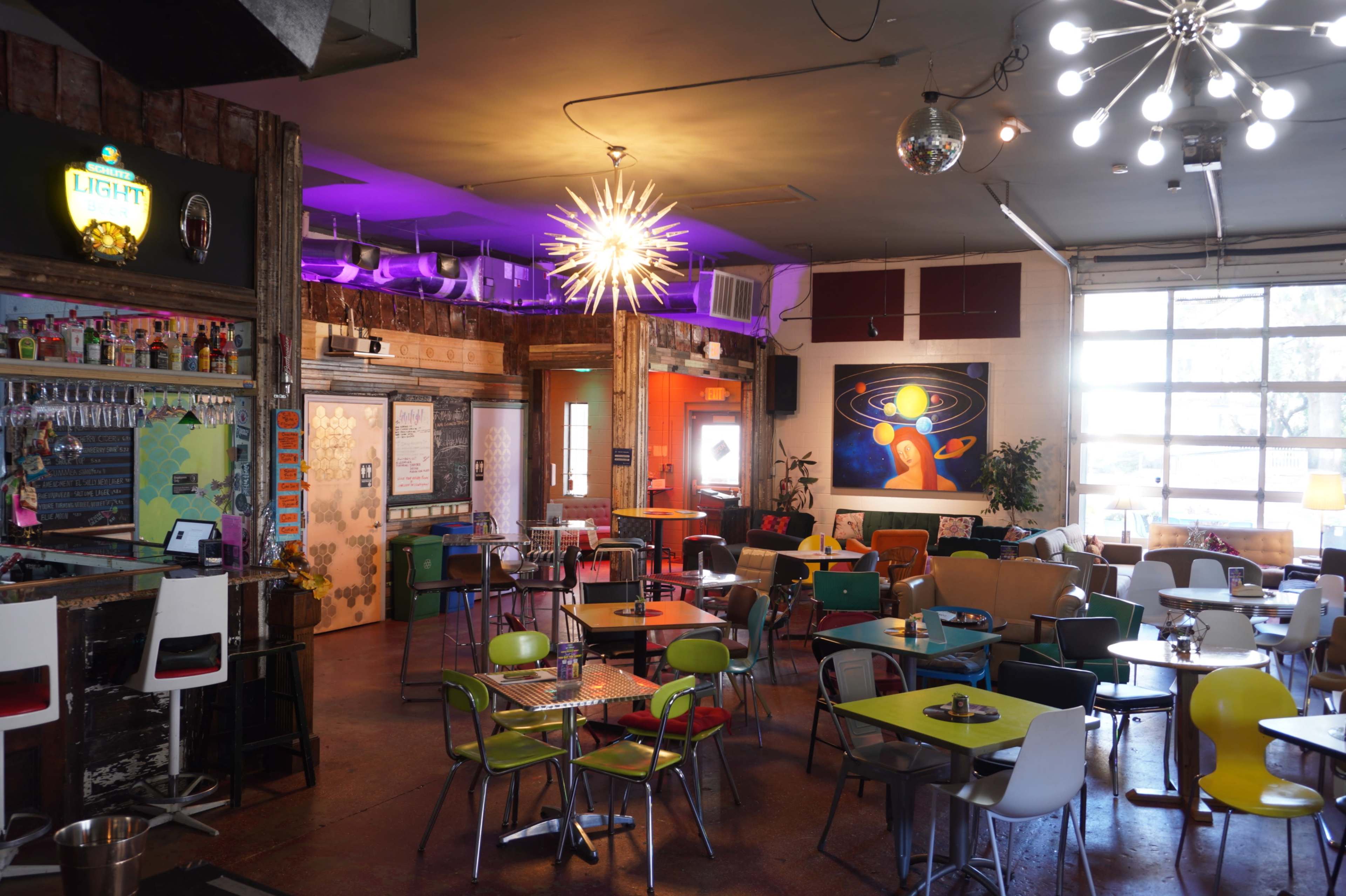 A vibrant café interior features colorful seating arrangements, a bar with various beverages, and eclectic lighting fixtures.