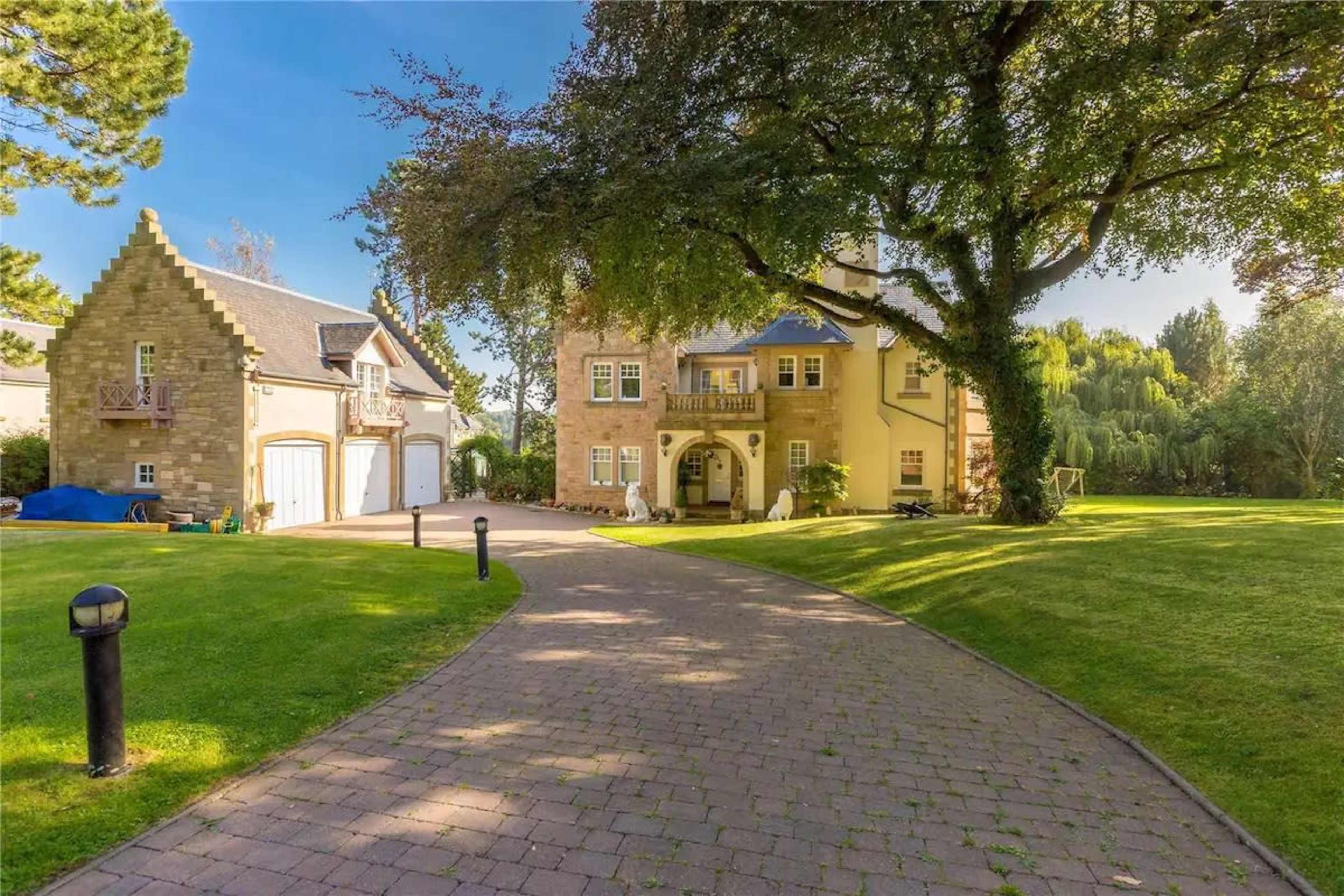A large house with a tower is set alongside a two-car garage, surrounded by manicured lawns and trees.