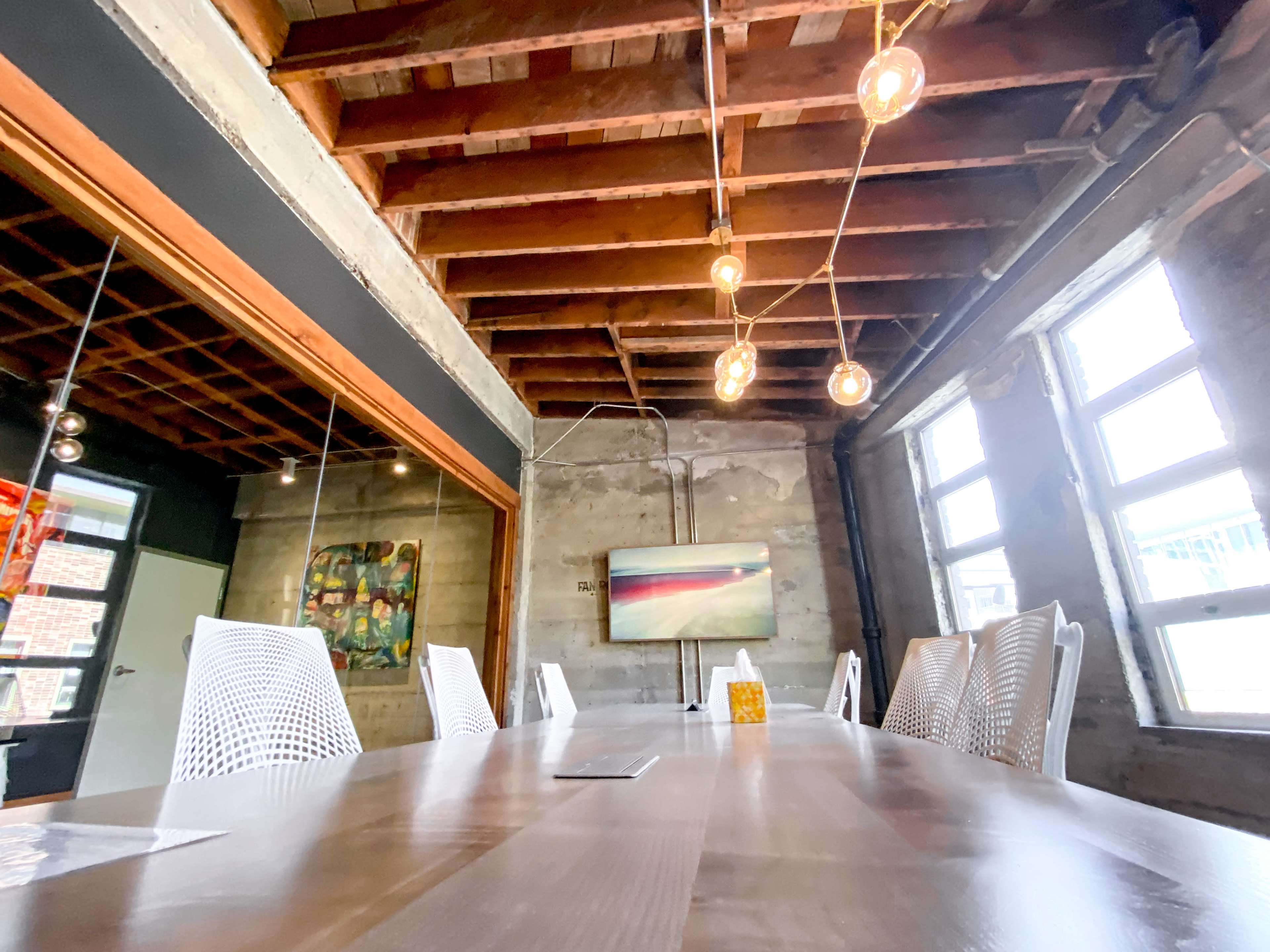 A long wooden table surrounded by white chairs is set in a room with exposed wooden beams and concrete walls, featuring large windows and a mounted television.