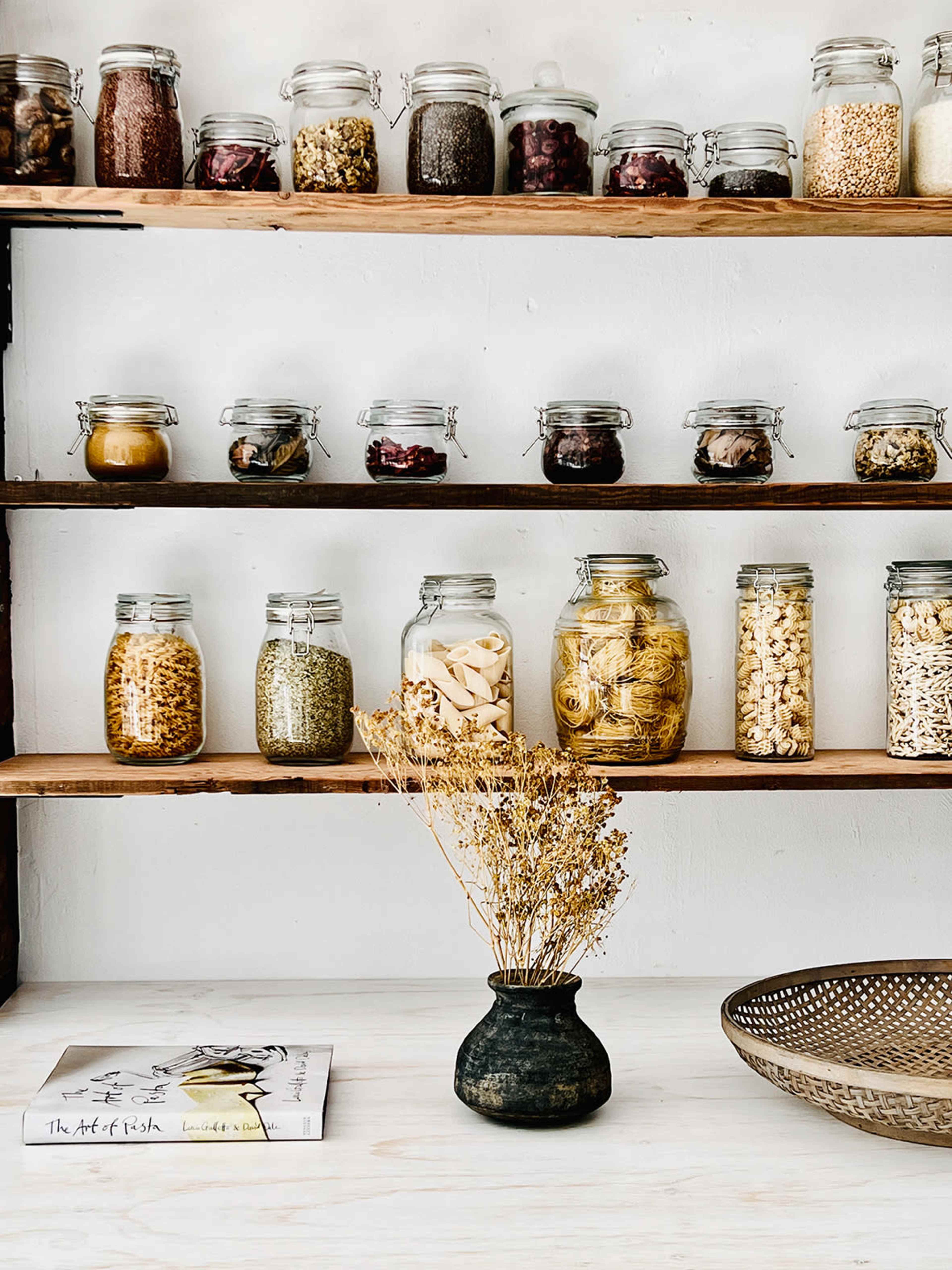 The image features a set of wooden shelves filled with glass jars containing various food items, accompanied by a small vase of dried flowers and a book on a white surface.