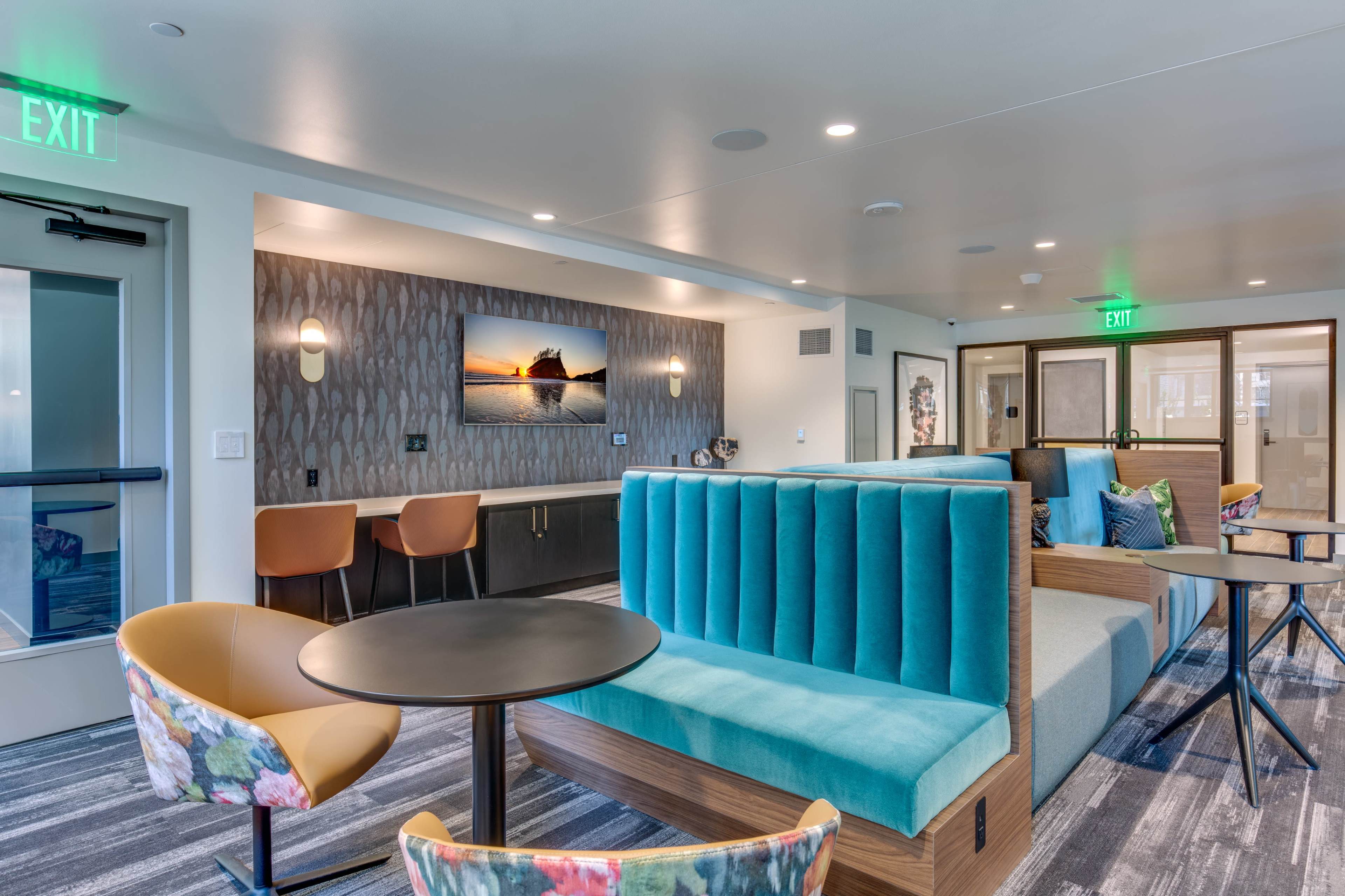 A modern lounge area featuring turquoise seating, a wall-mounted television displaying a sunset, and a mix of round and square tables.