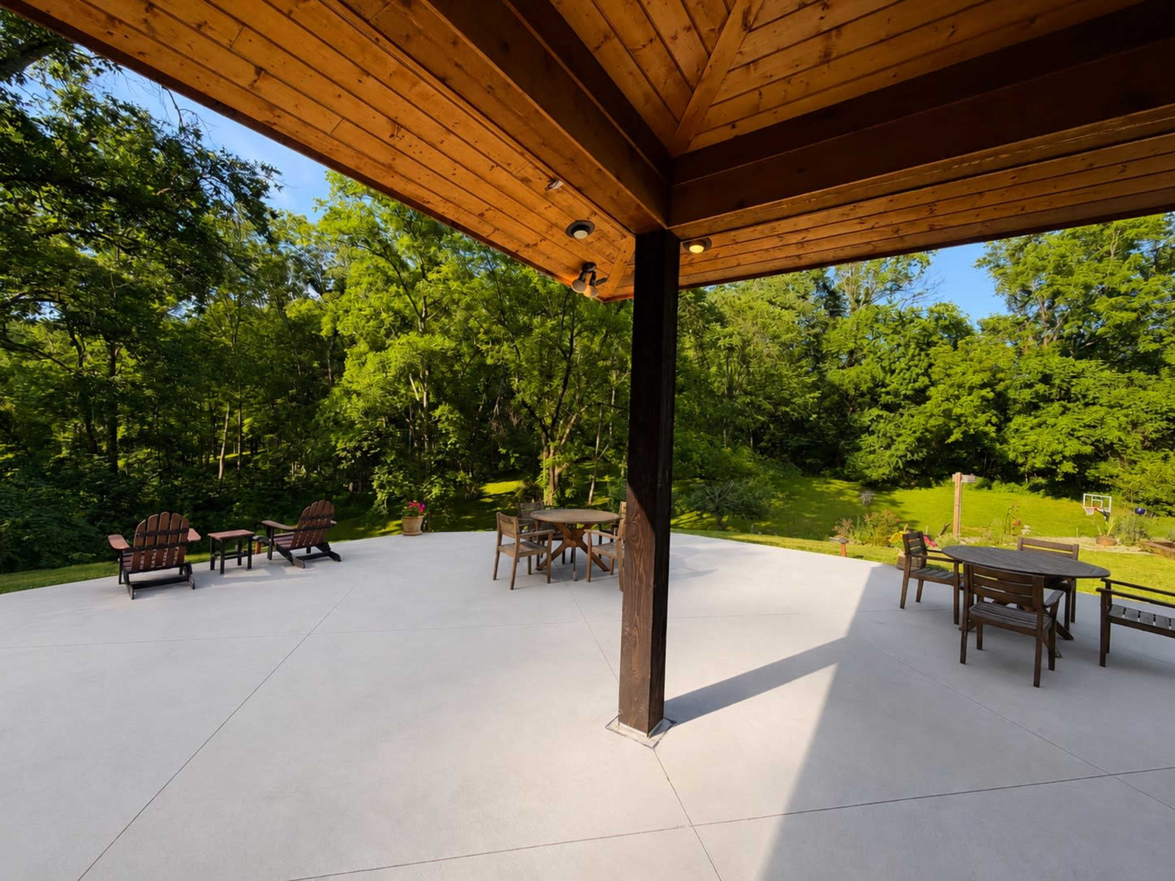 The image shows a modern outdoor patio area surrounded by lush trees and greenery, with a wooden ceiling overhead.