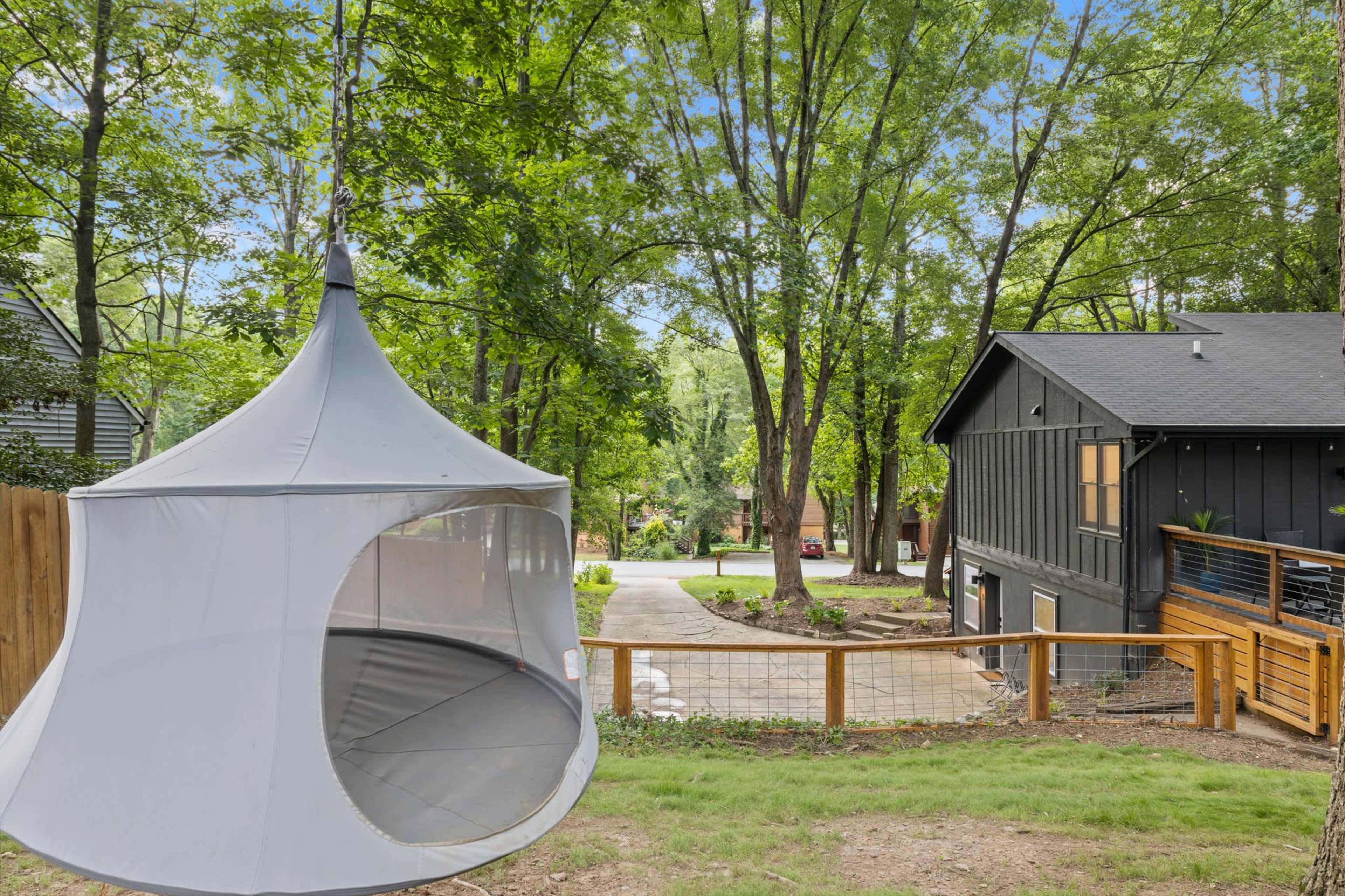 A backyard with a suspended tent-like structure and a modern house surrounded by trees, leading to a gravel driveway.