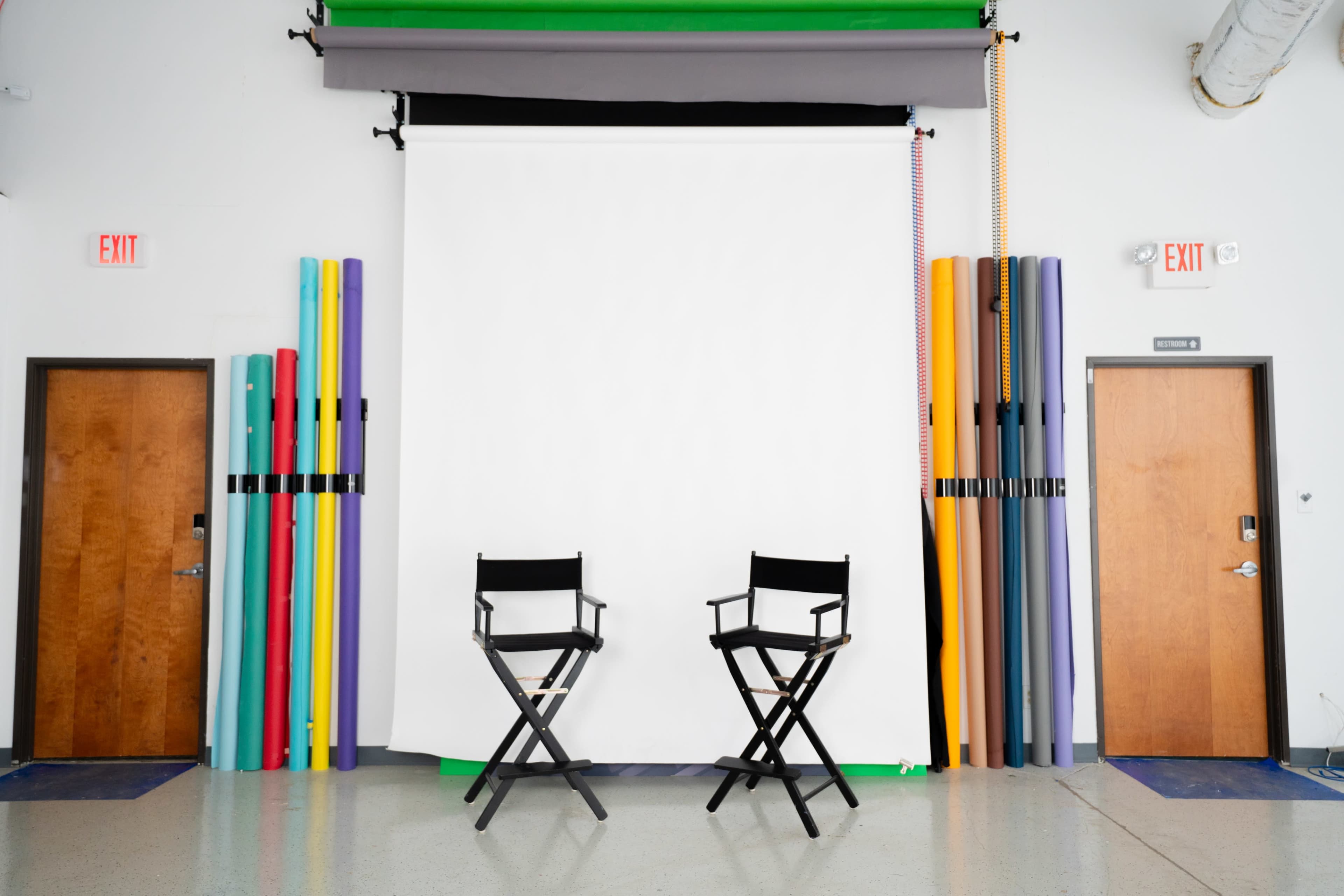 The image shows a photography studio setup featuring two black director's chairs, positioned in front of a large white backdrop, with colorful rolls of backdrop paper stacked beside the walls.