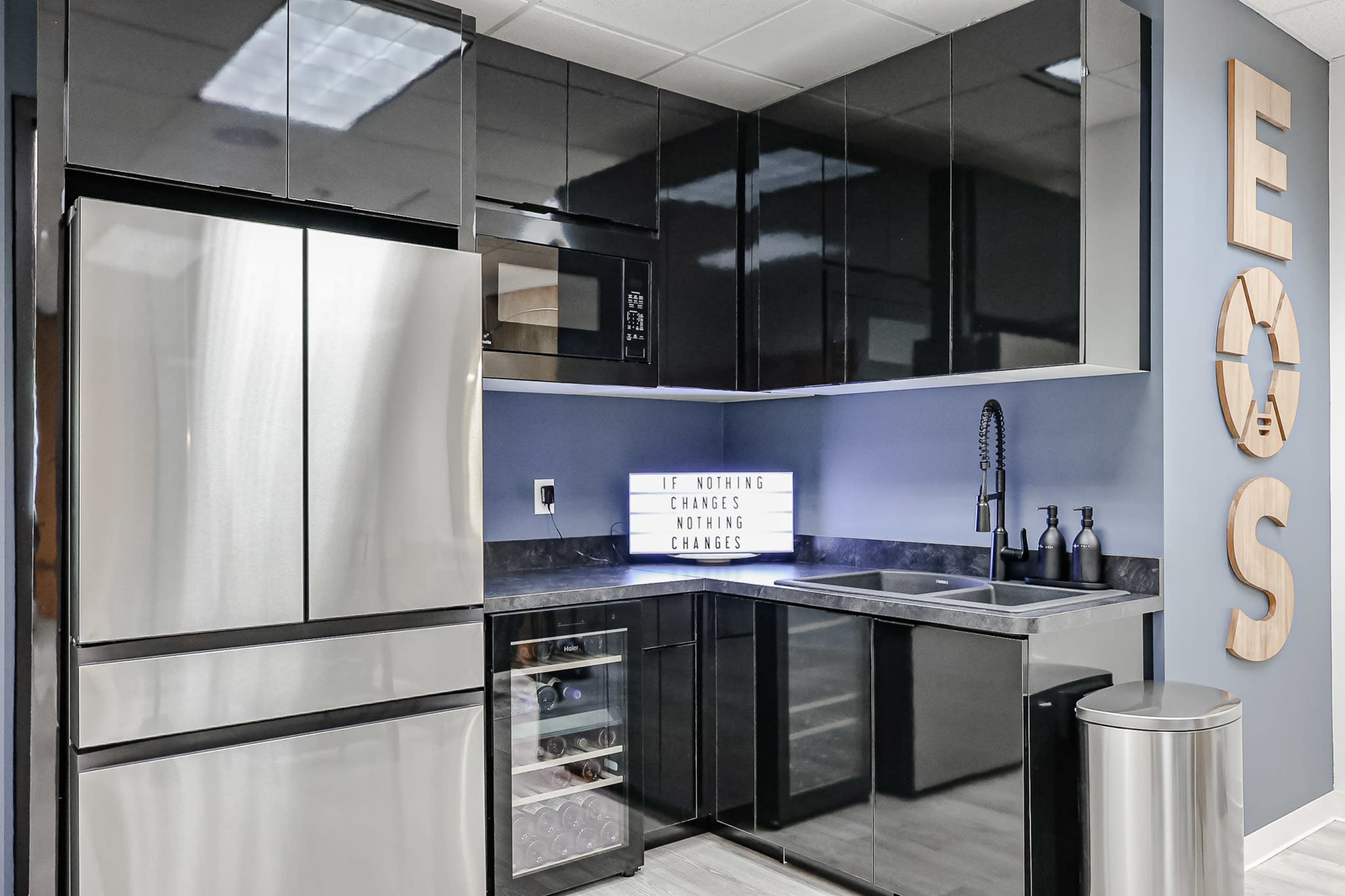 A modern kitchen features black cabinetry, stainless steel appliances, and a lighted sign with motivational text.