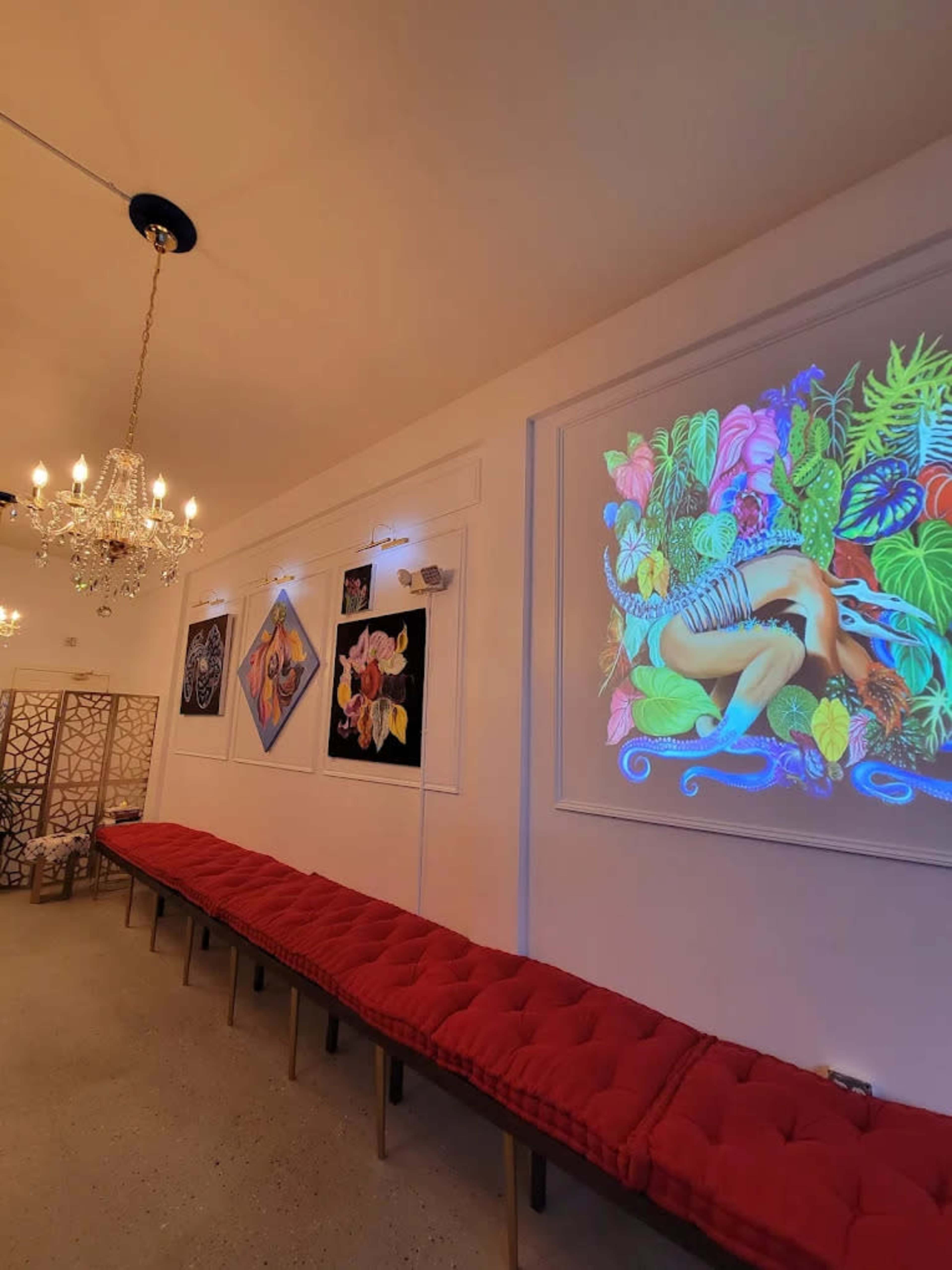 The image shows an art gallery interior with a long red bench, a large colorful artwork projected on the wall, and ornate chandeliers hanging from the ceiling.