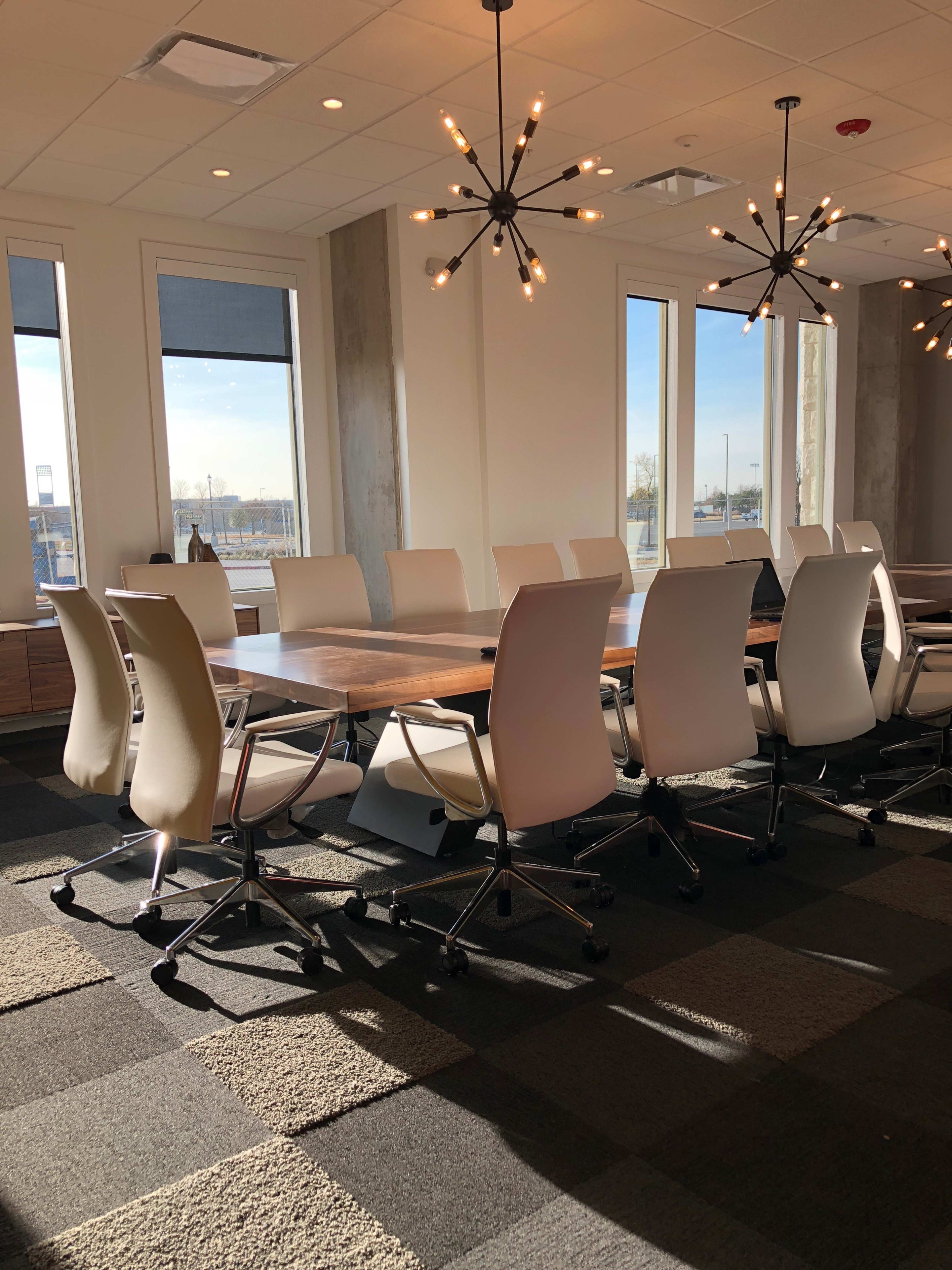 A conference room features a large table surrounded by ergonomic chairs and modern light fixtures, with windows providing natural light.