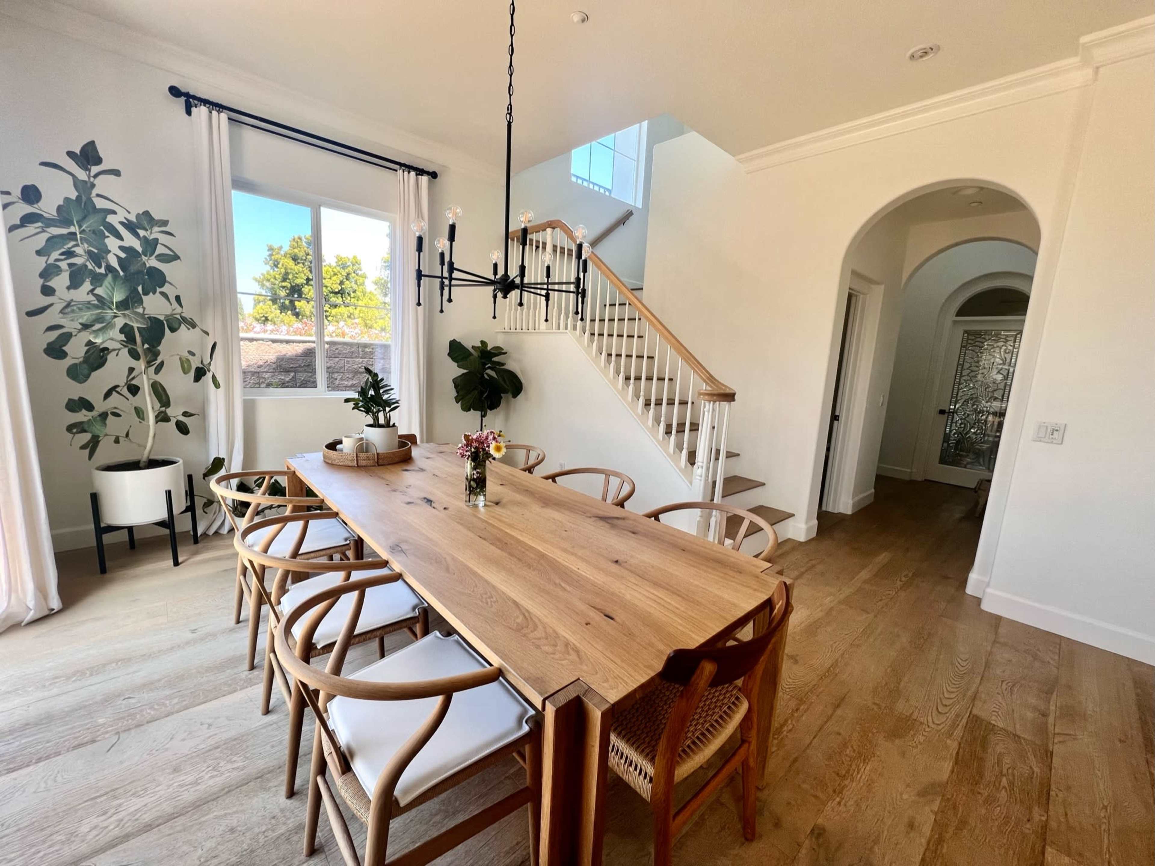 A dining area features a wooden table surrounded by chairs, with large windows and a staircase visible in the background.
