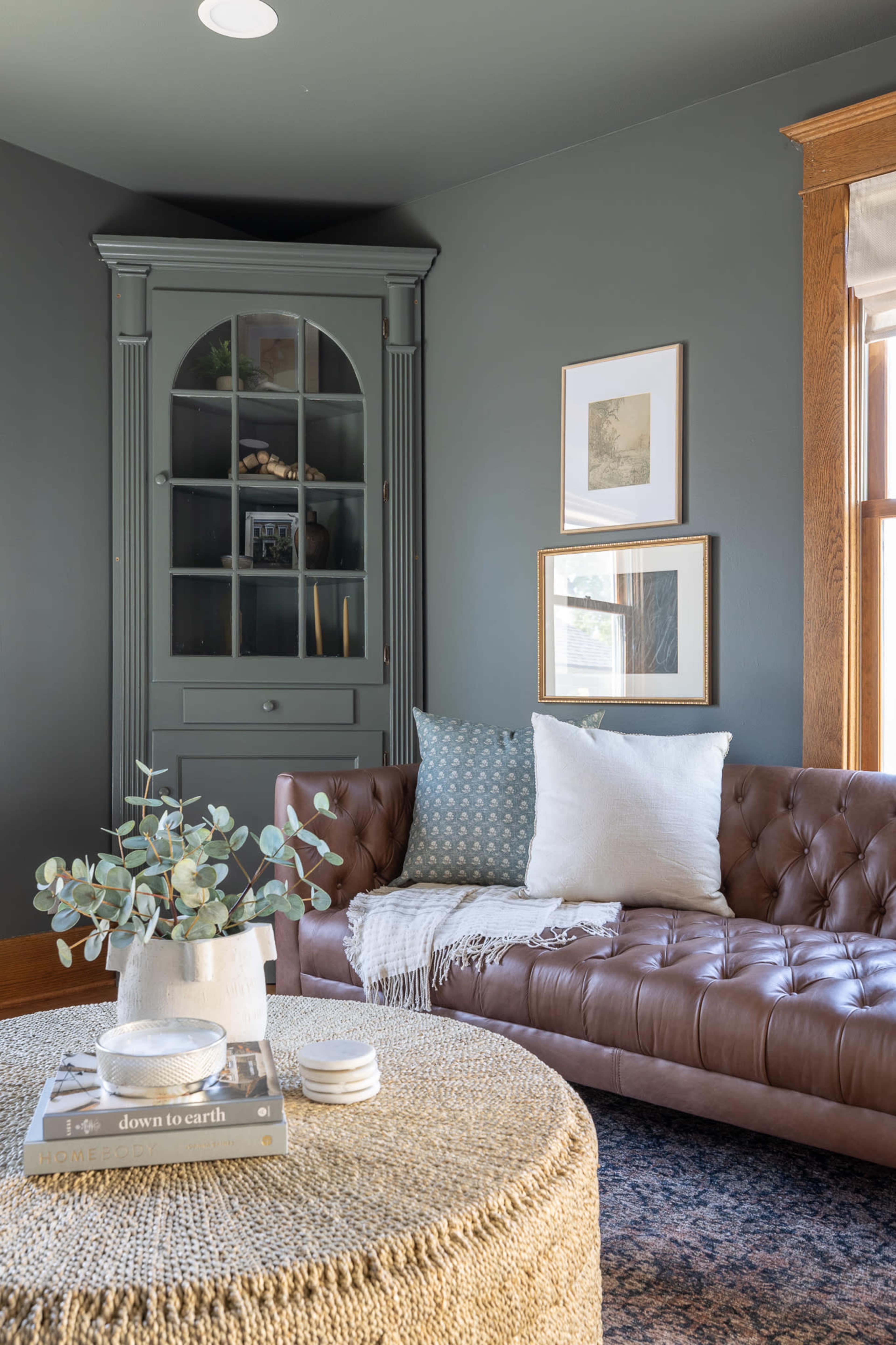 A cozy living room with a brown leather sofa, a round woven coffee table, and a green cabinet in the corner against dark walls.