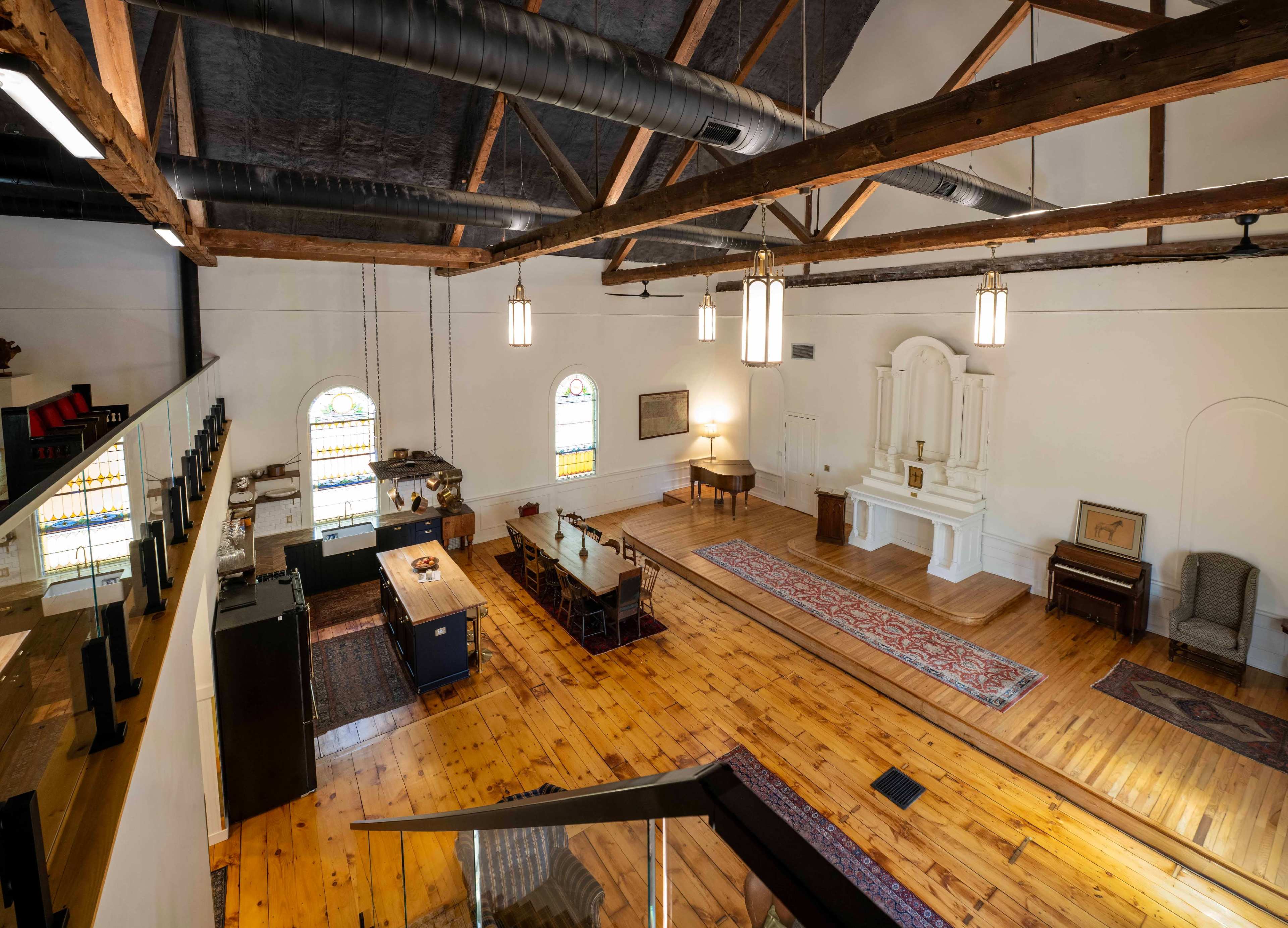 The image shows the interior of a spacious room with wooden flooring, high ceilings, and a long table set for dining, complemented by various seating areas and decorative windows.