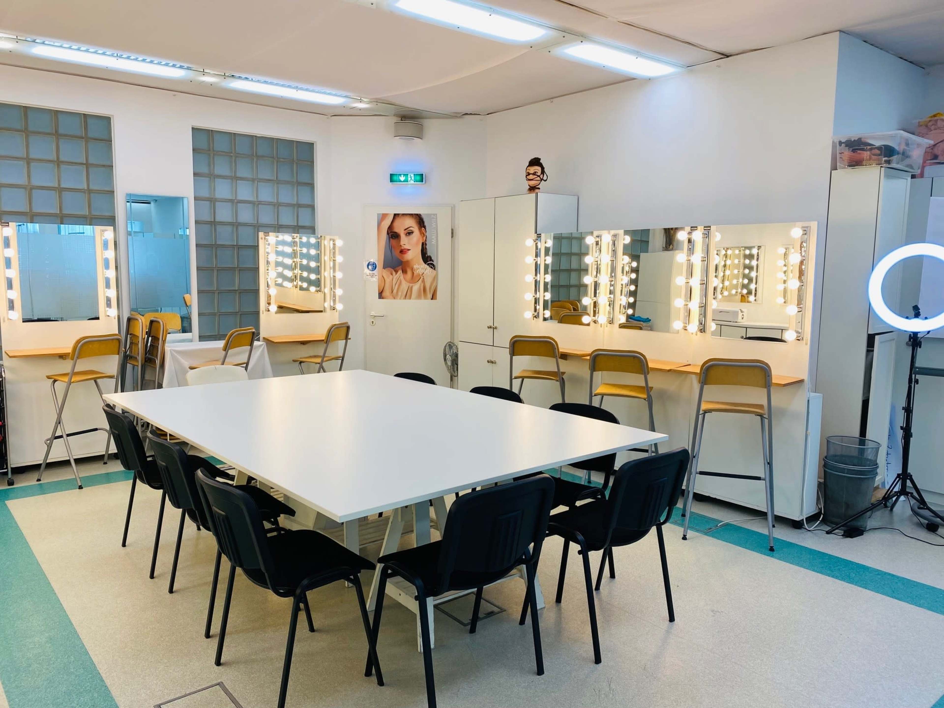 A well-lit dressing room with a large table and several chairs, featuring illuminated mirrors along the walls.