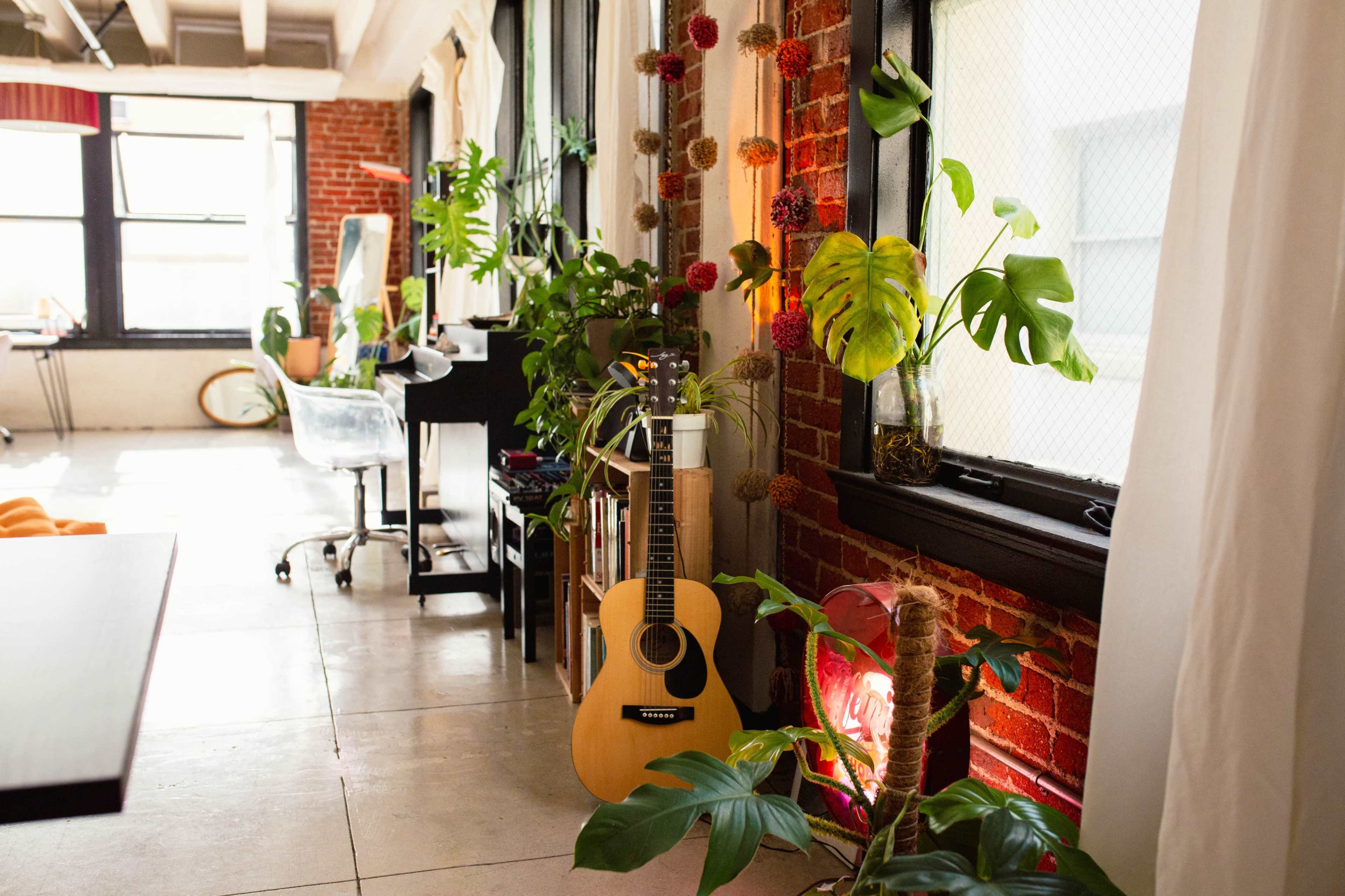 The image shows a bright, airy room featuring a wooden guitar leaning against a brick wall, surrounded by assorted indoor plants and large windows.