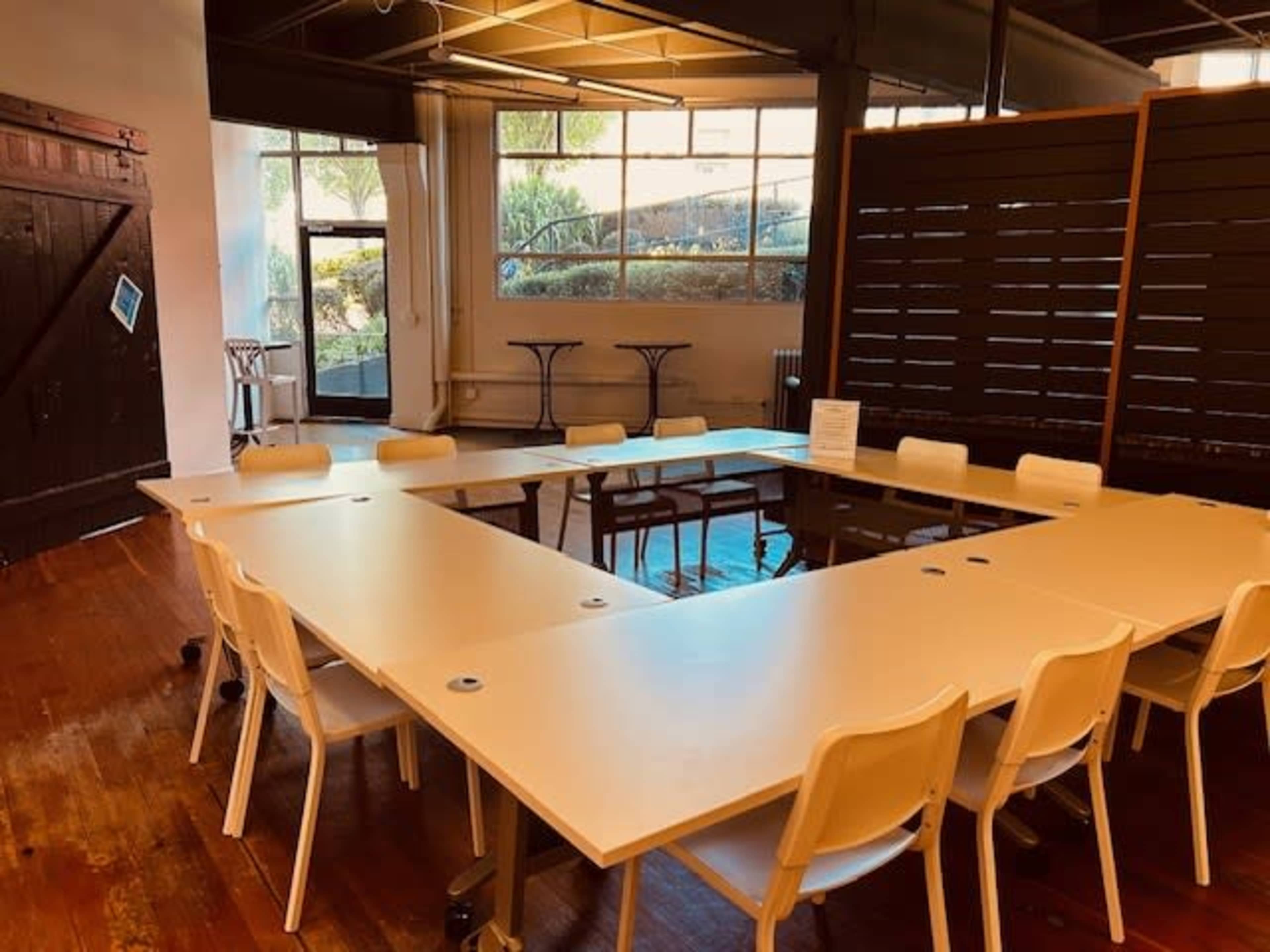 A meeting room features a U-shaped arrangement of white tables and chairs with large windows allowing natural light.