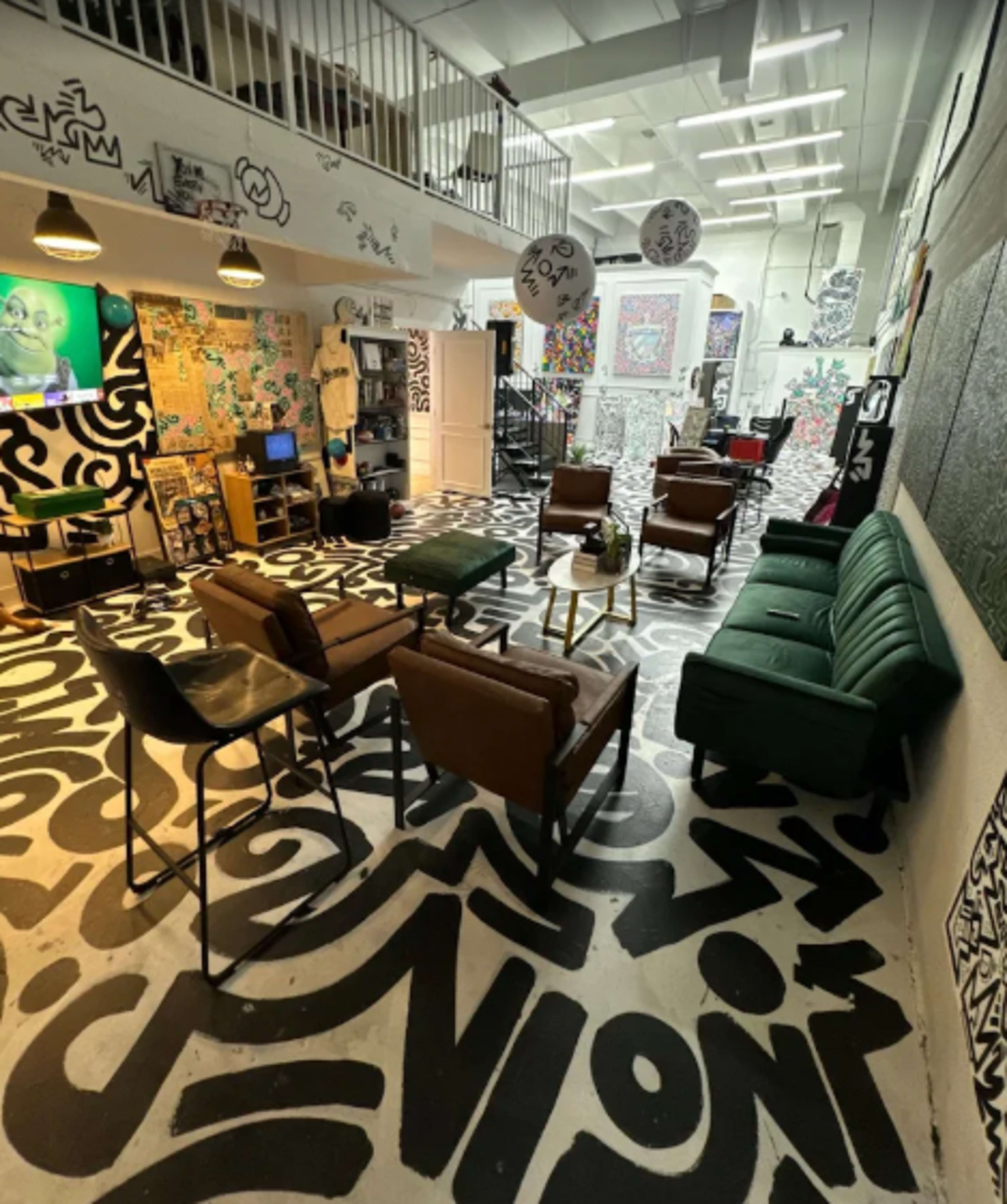 The image shows a vibrant lounge area with eclectic art-covered walls, patterned flooring, and a mix of modern furniture, including chairs and tables.
